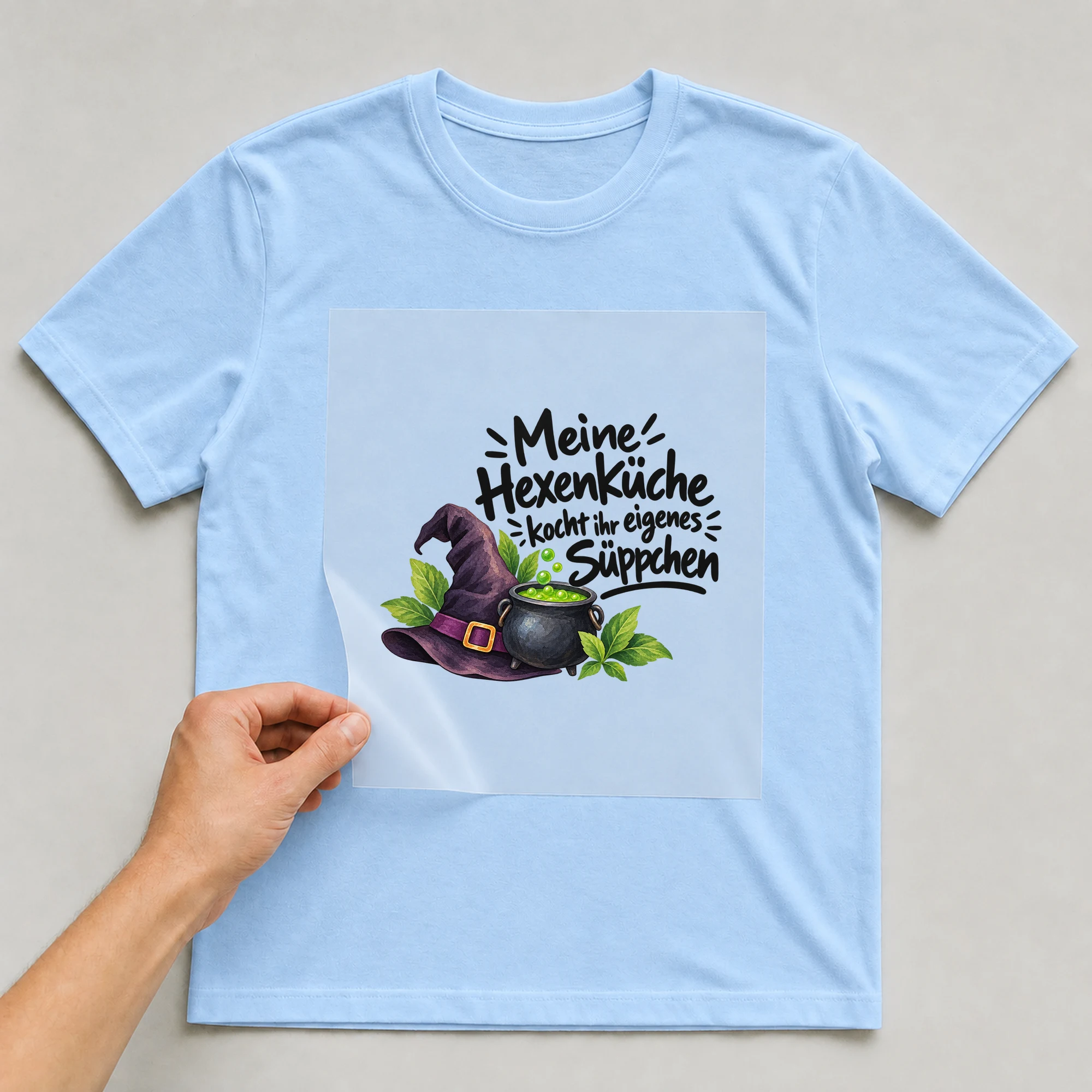 Bügelbild „Meine Hexenküche kocht ihr eigenes Süppchen“ auf babyblauem T-Shirt, Folie wird aufgelegt