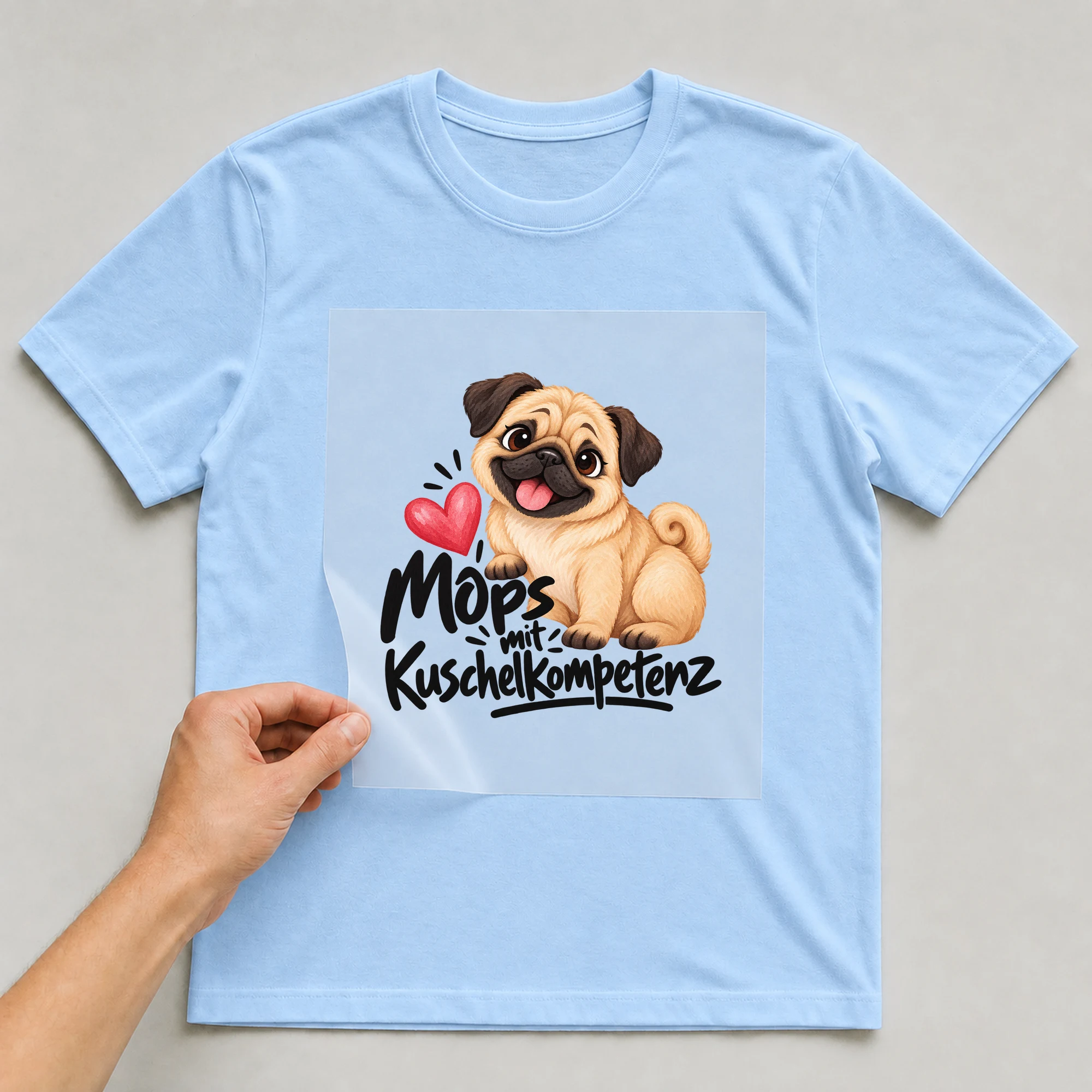 Bügelbild „Mops mit Kuschelkompetenz“ auf hellblauem T-Shirt, Folie wird aufgelegt