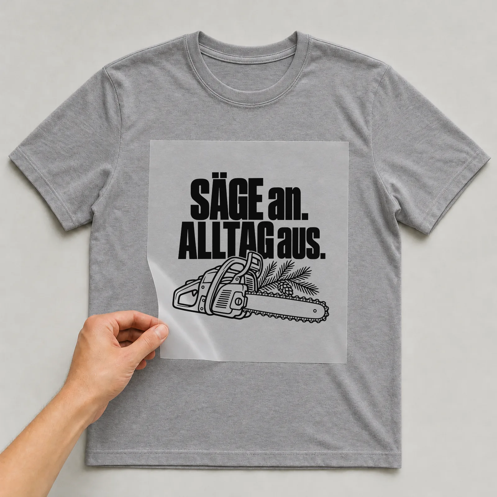 Bügelbild „Säge an. Alltag aus.“ auf grauem T-Shirt, Folie wird aufgelegt