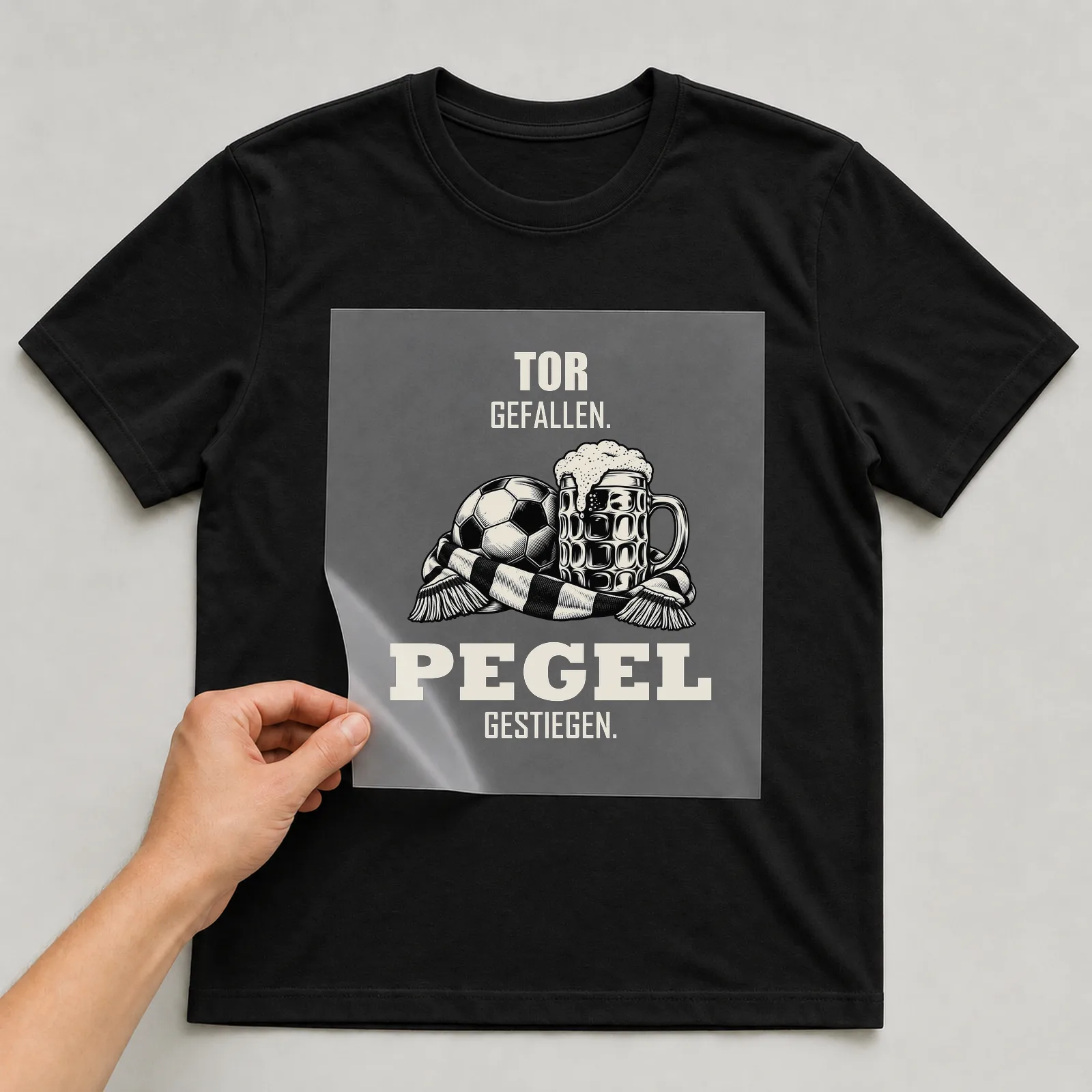 Bügelbild „Tor gefallen. Pegel gestiegen.“ auf schwarzem T-Shirt, Folie wird aufgelegt
