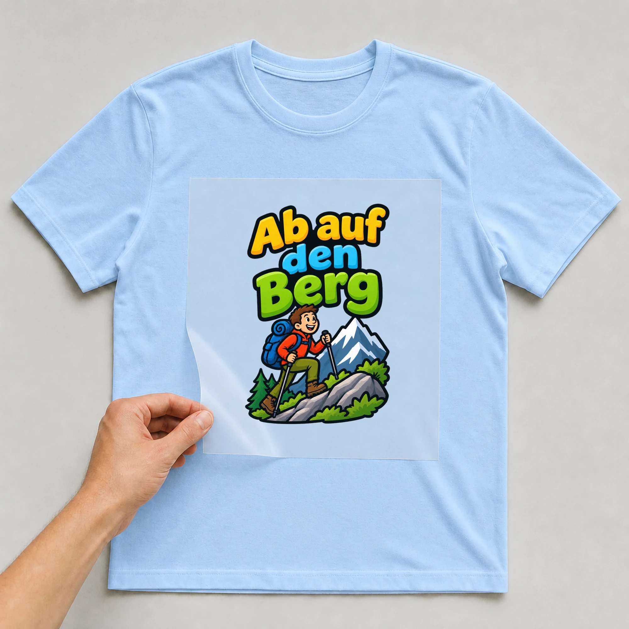 Bügelbild „Ab auf den berg“ auf babyblauem T-Shirt, Folie wird aufgelegt