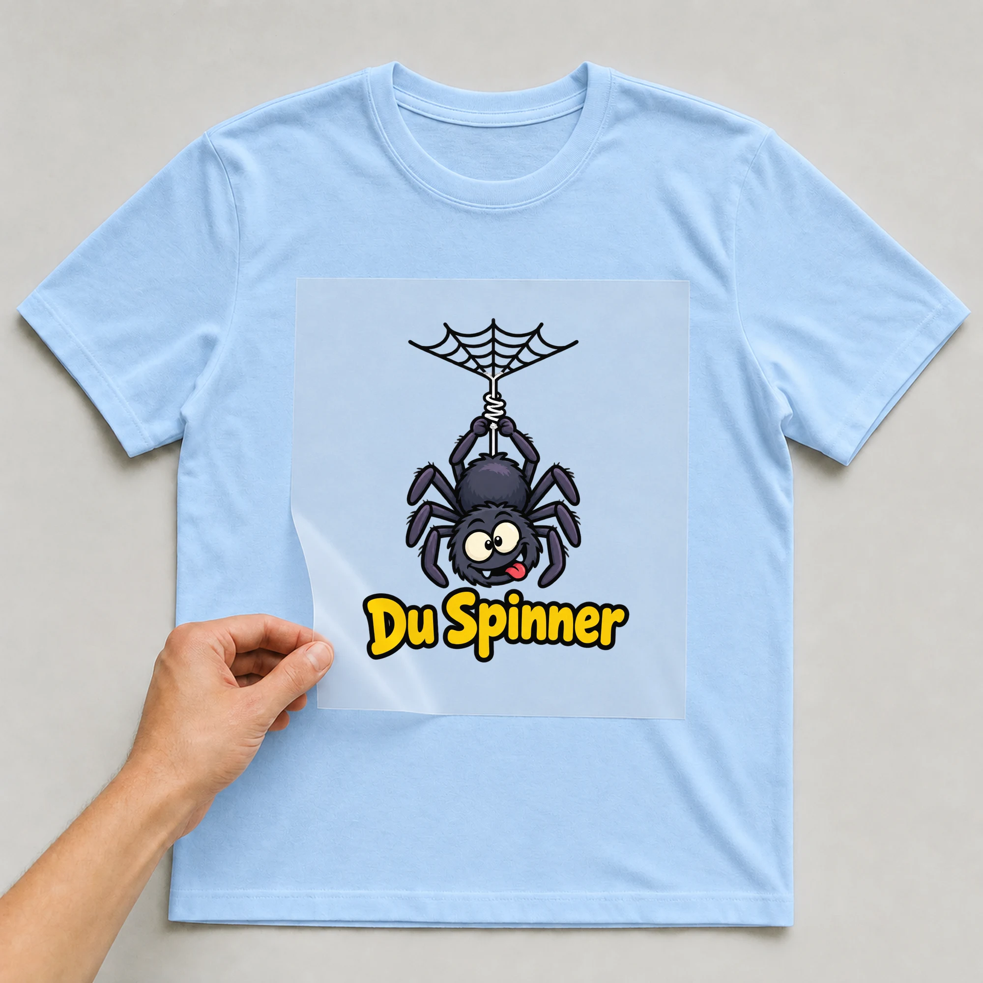 Bügelbild „Du Spinner“ auf babyblauem T-Shirt, Folie wird aufgelegt