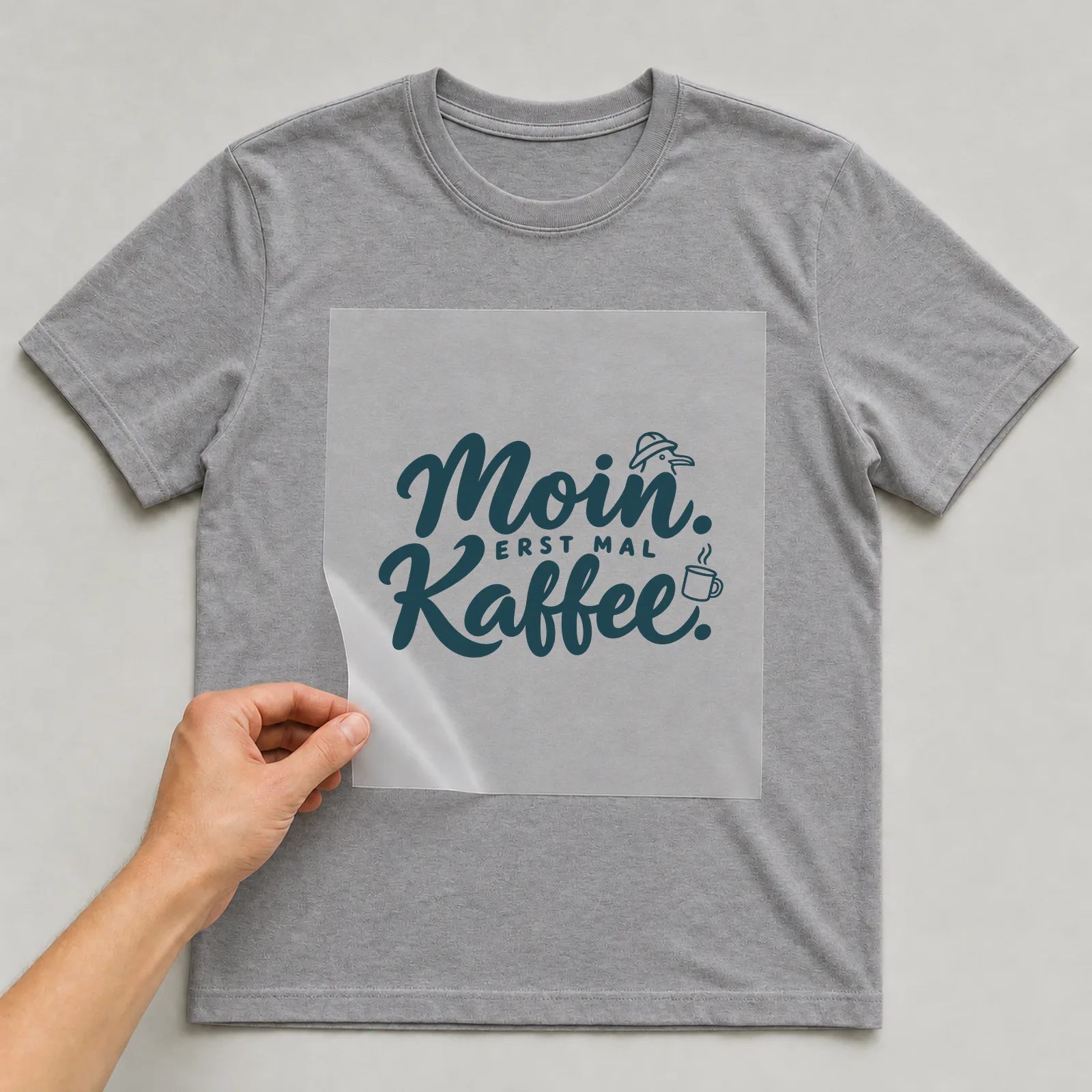 Bügelbild „Moin. Erst mal Kaffee.“ (Schrift Konturen, Nr. 2) auf grauem T-Shirt, Folie wird aufgelegt