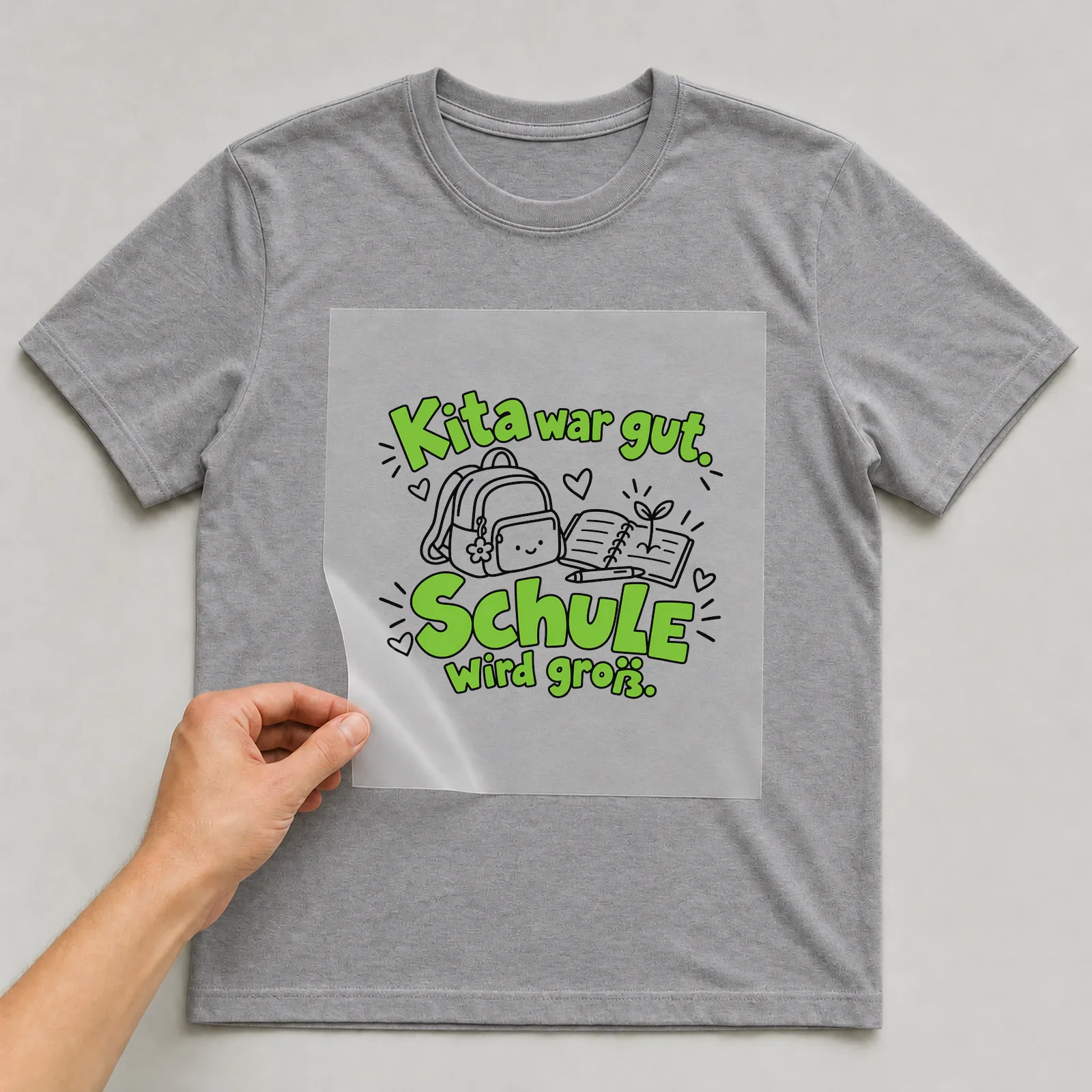 Bügelbild „Kita war gut. Schule wird groß.“ auf grauem T-Shirt, Folie wird aufgelegt