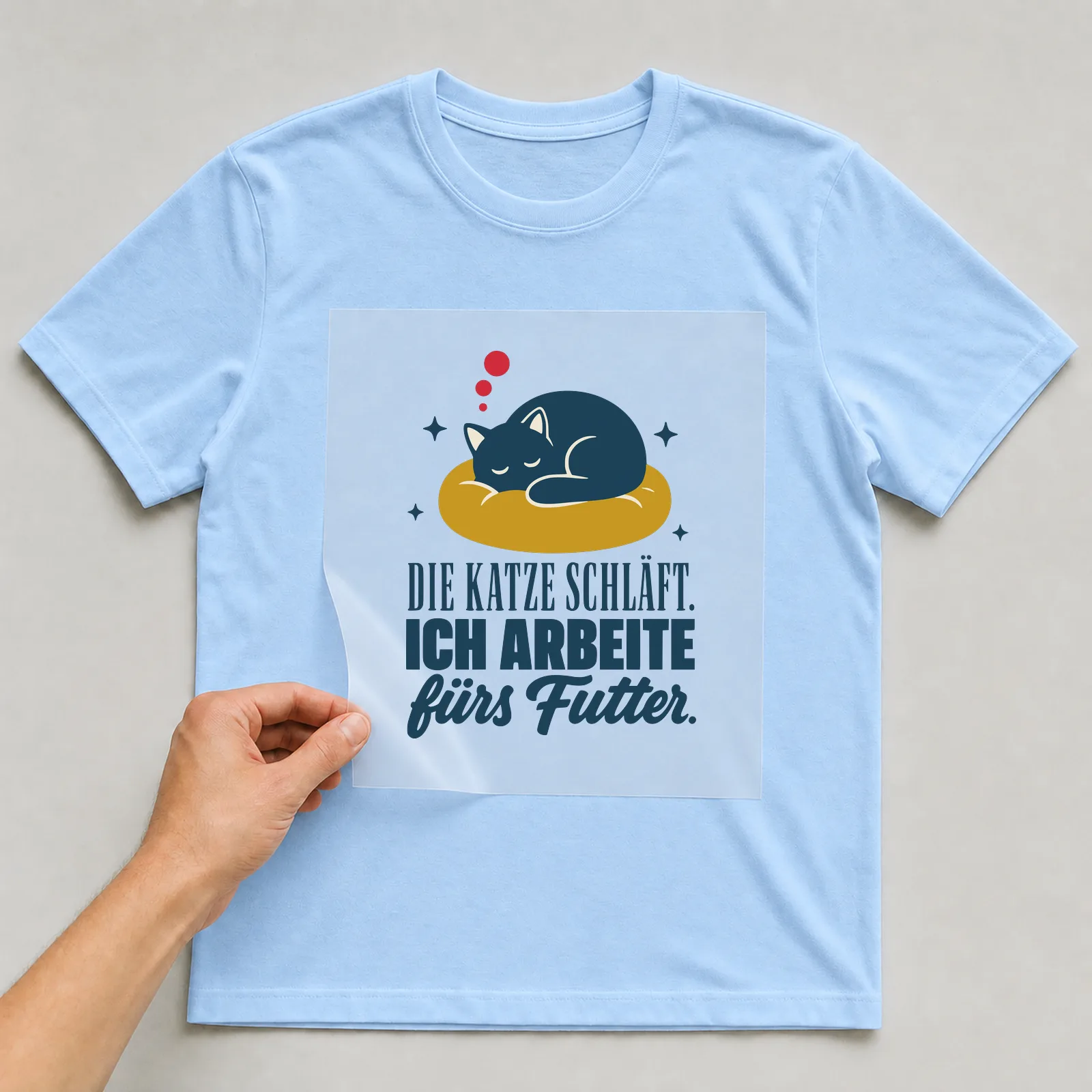 Bügelbild Die Katze schläft. Ich arbeite fürs Futter. auf hellblauem T-Shirt, Folie wird aufgelegt