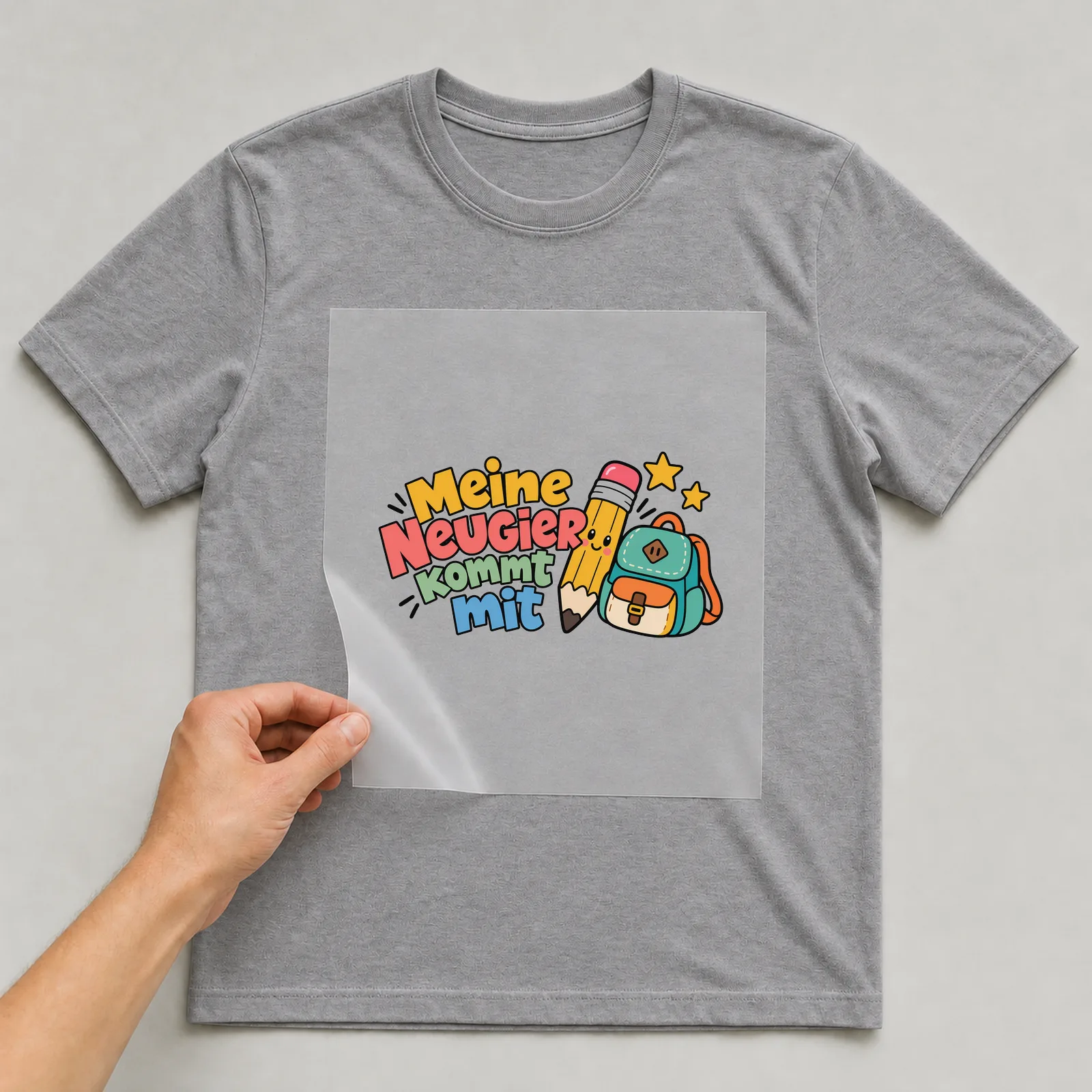 Bügelbild „Meine Neugier kommt mit“ auf grauem T-Shirt, Folie wird aufgelegt