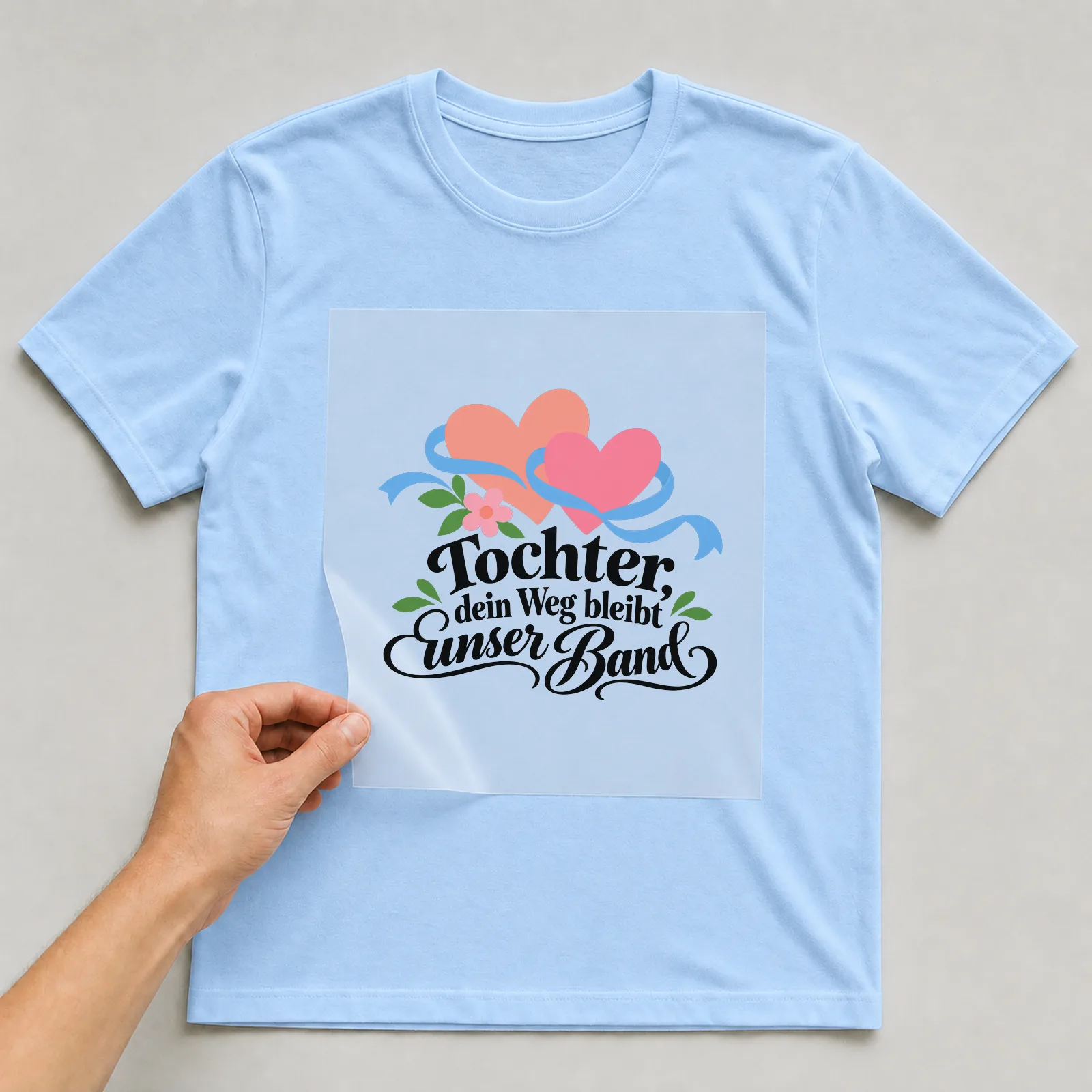 Bügelbild „Tochter, dein Weg bleibt unser Band“ auf hellblauem T-Shirt, Folie wird aufgelegt