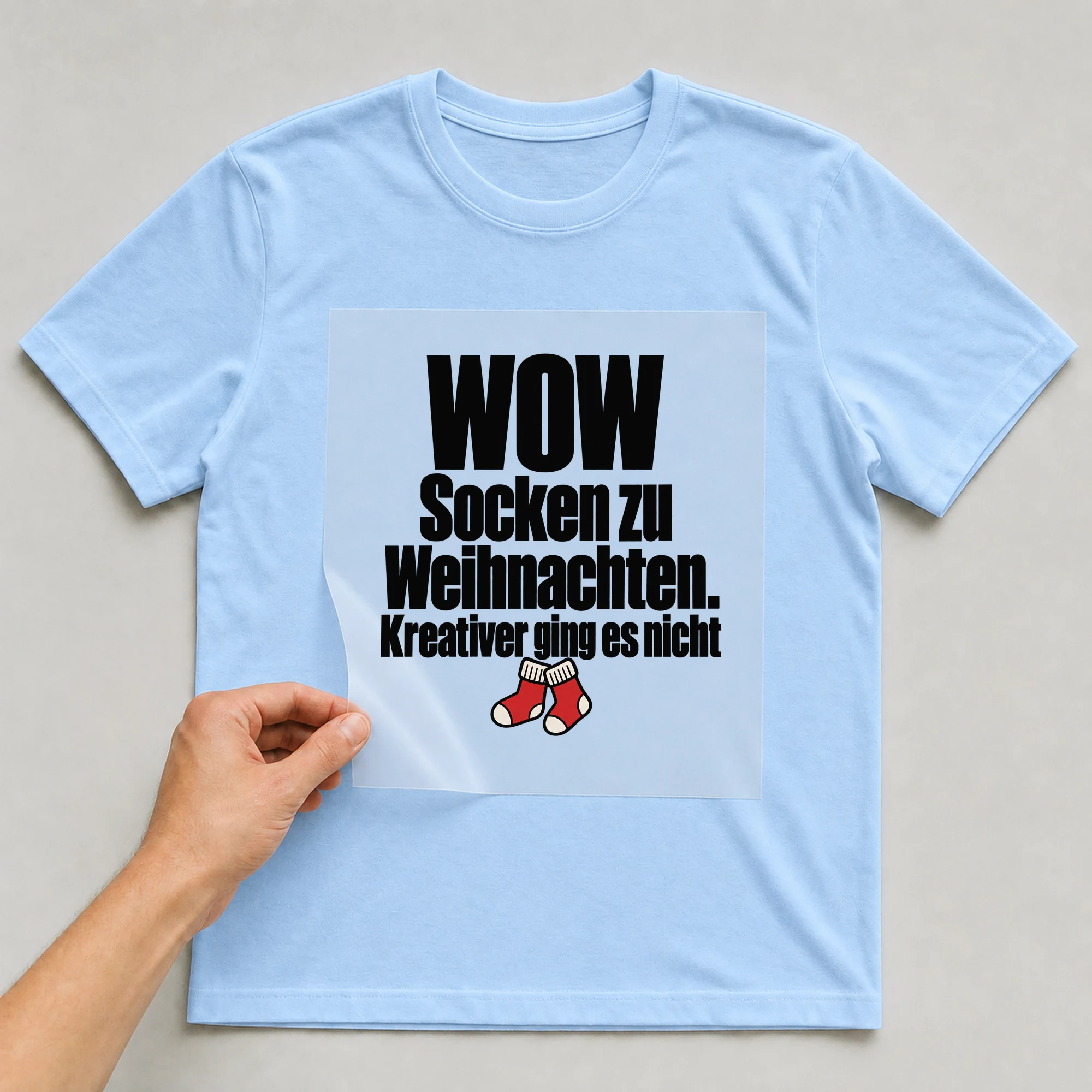 Bügelbild „WOW Socken zu Weihnachten. Kreativer ging es nicht“ auf babyblauem T-Shirt, Folie wird aufgelegt