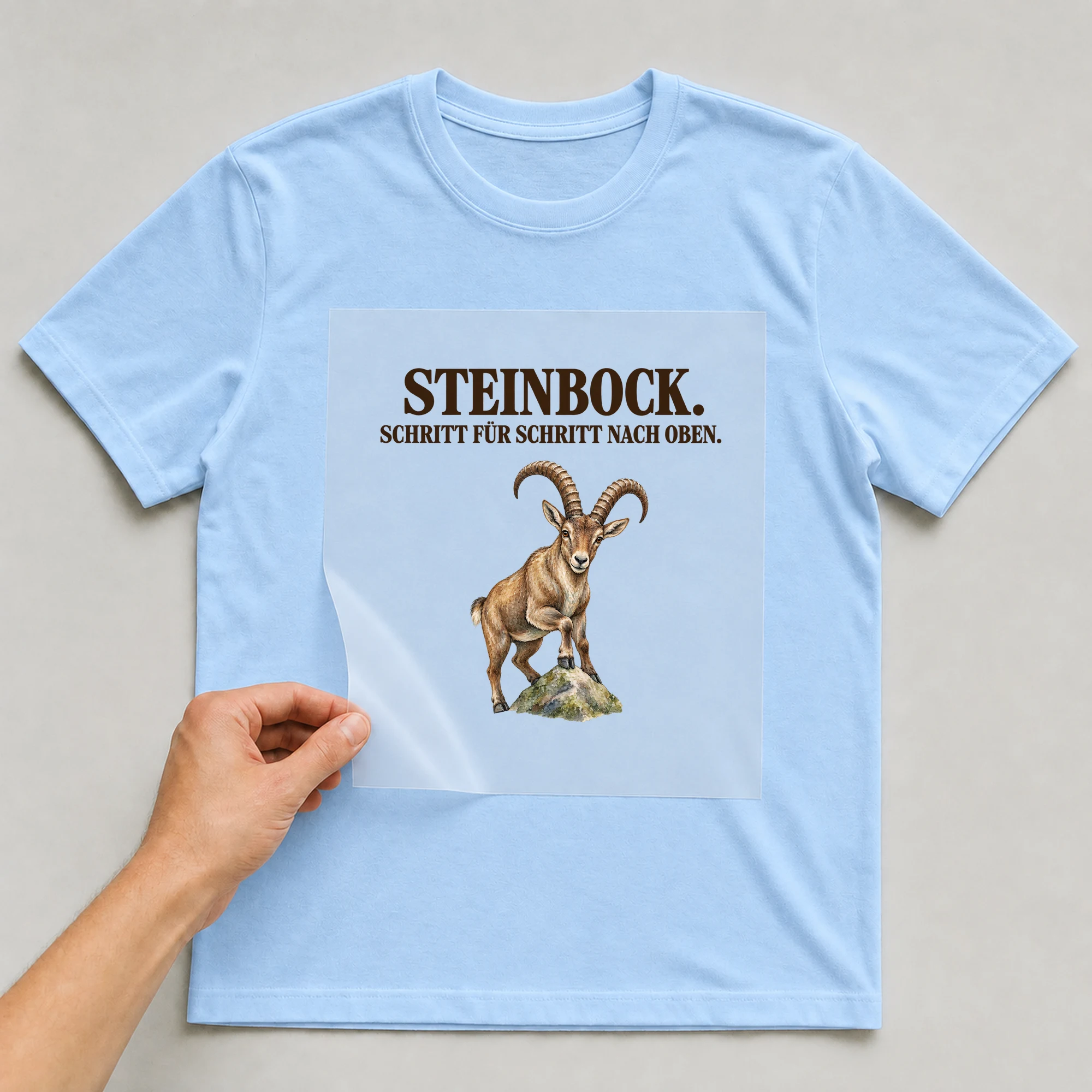 Bügelbild „STEINBOCK. SCHRITT FÜR SCHRITT NACH OBEN.“ auf babyblauem T-Shirt, Folie wird aufgelegt
