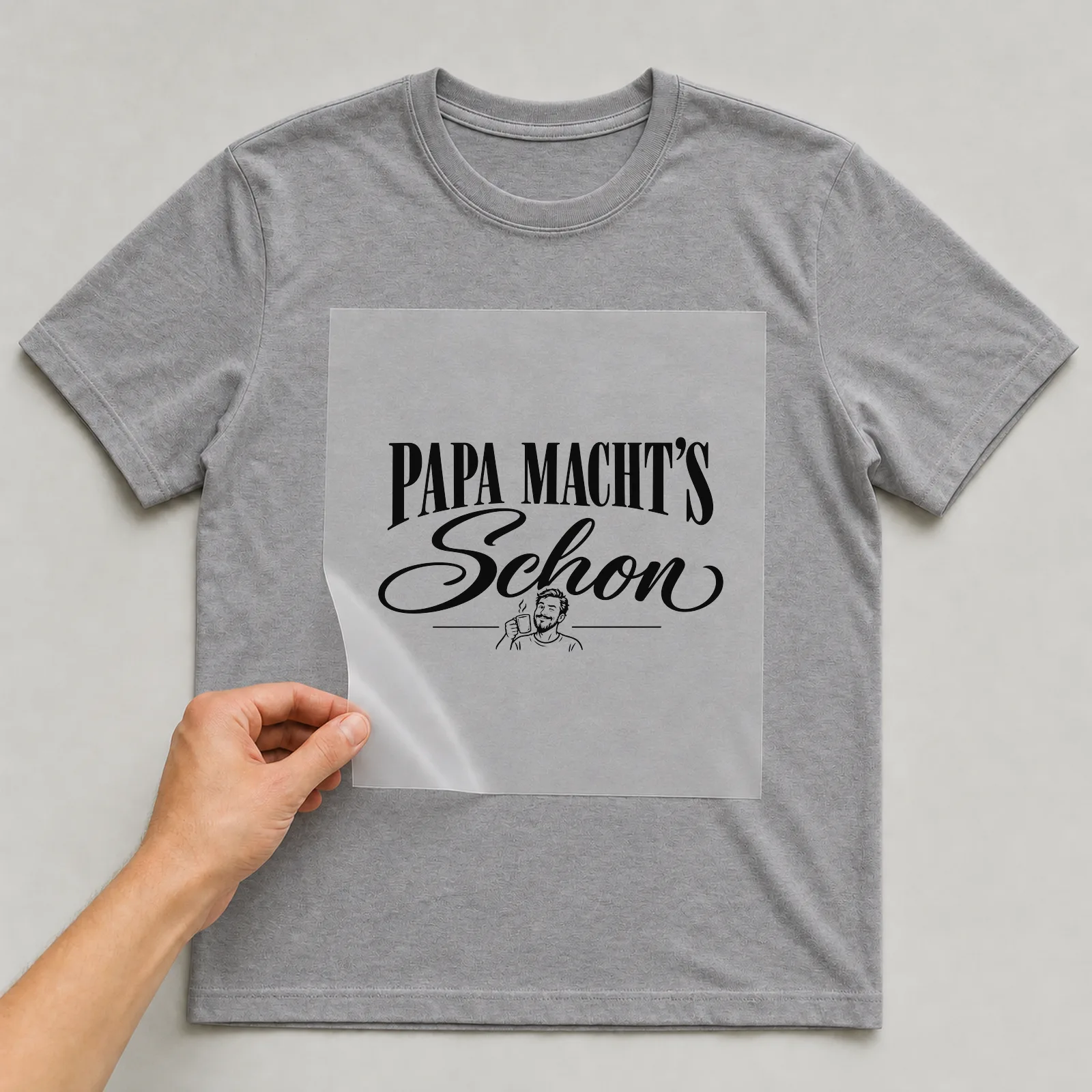 Bügelbild „Papa macht’s schon“ auf grauem T-Shirt, Folie wird aufgelegt