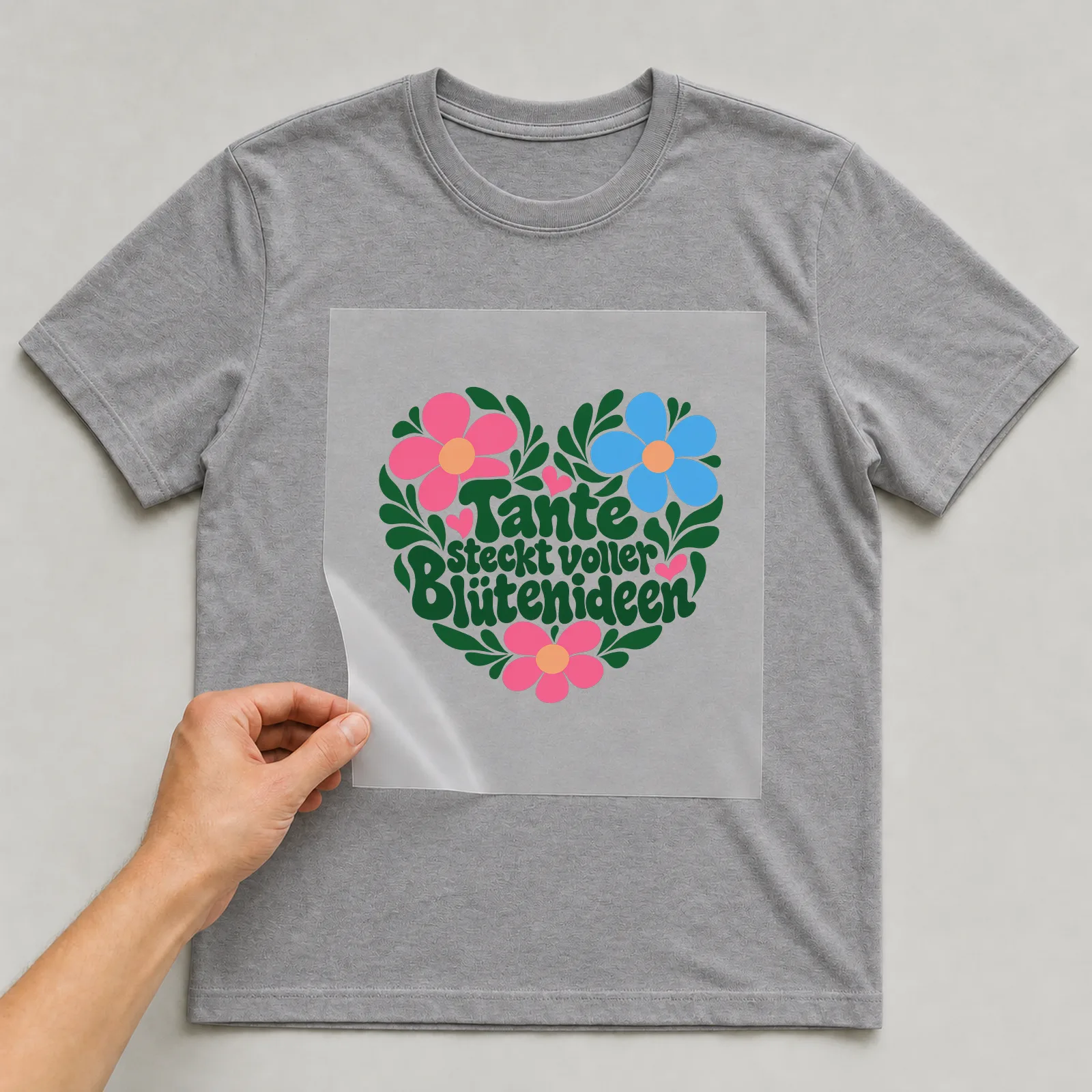 Bügelbild „Tante steckt voller Blütenideen“ auf grauem T-Shirt, Folie wird aufgelegt