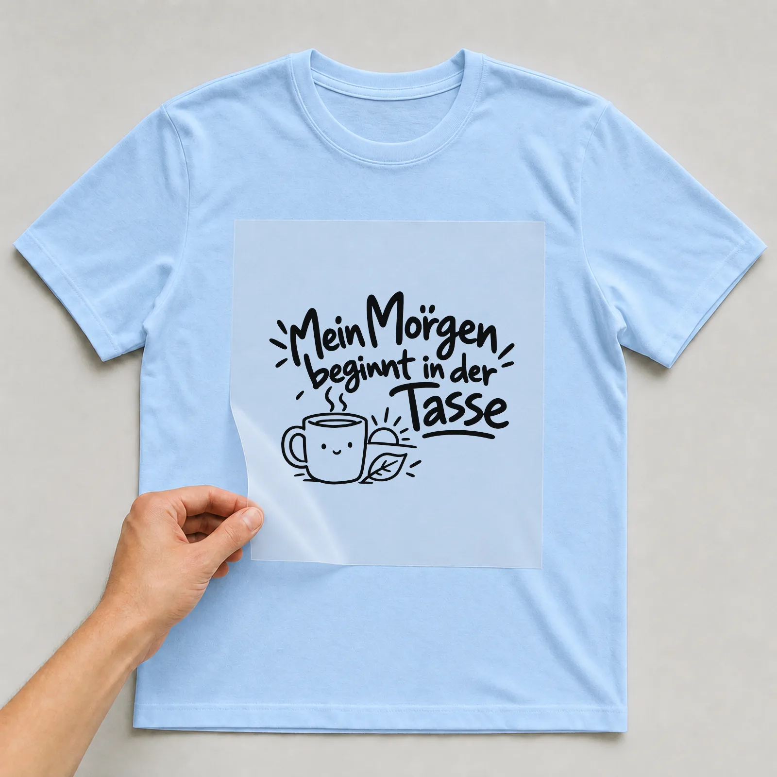 Bügelbild „Mein Morgen beginnt in der Tasse“ auf hellblauem T-Shirt, Folie wird aufgelegt
