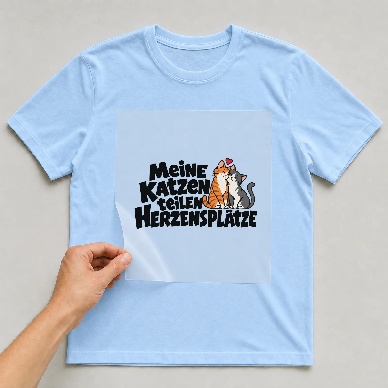 Bügelbild „Meine Katzen teilen Herzensplätze“ auf hellblauem T-Shirt, Folie wird aufgelegt