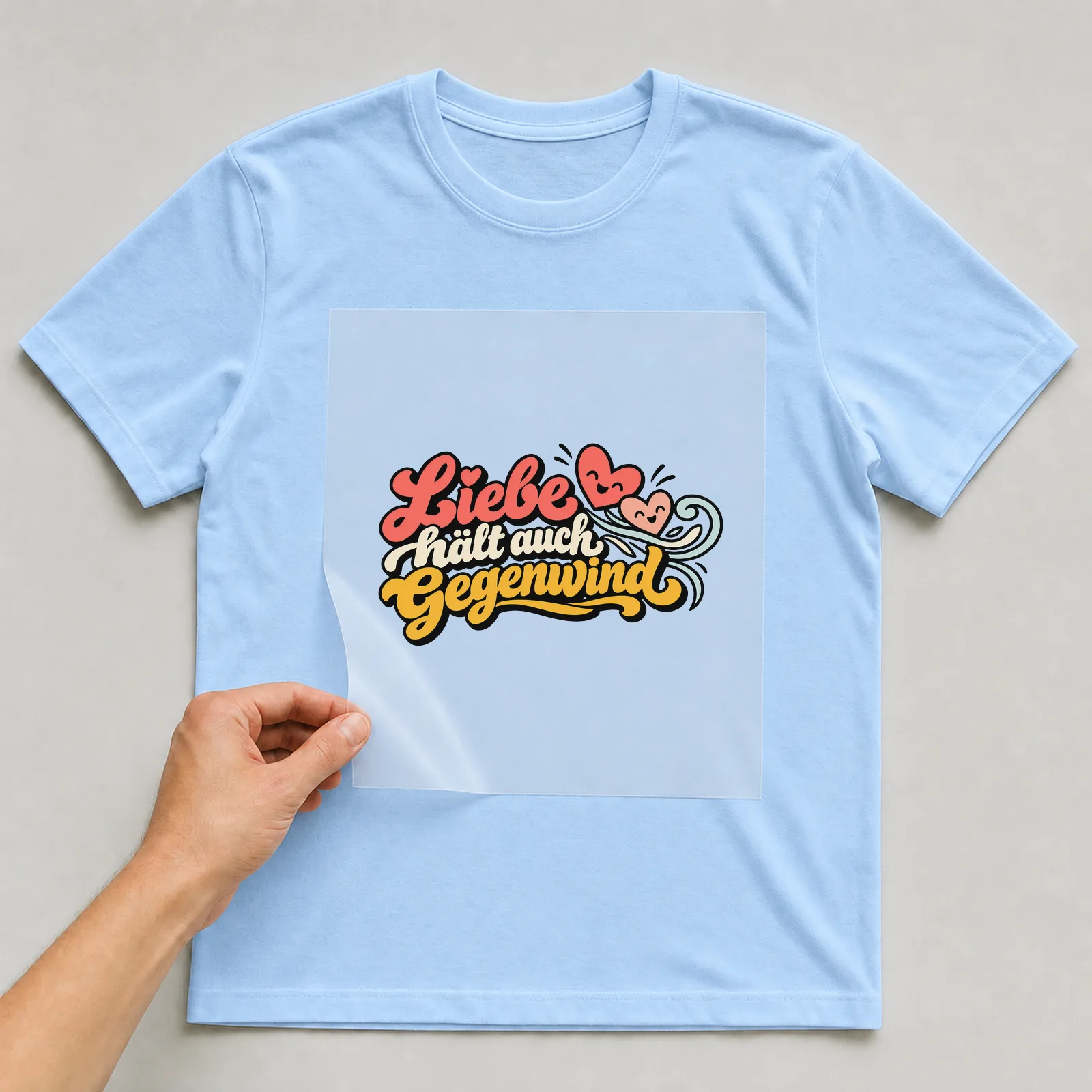 Bügelbild „Liebe hält auch Gegenwind“ auf hellblauem T-Shirt, Folie wird aufgelegt