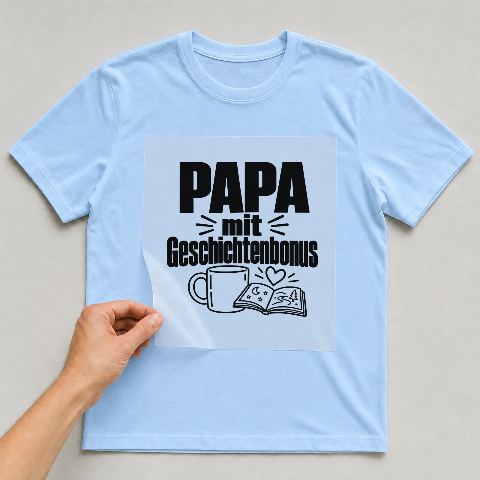 Bügelbild „Papa mit Geschichtenbonus“ auf hellblauem T-Shirt, Folie wird aufgelegt