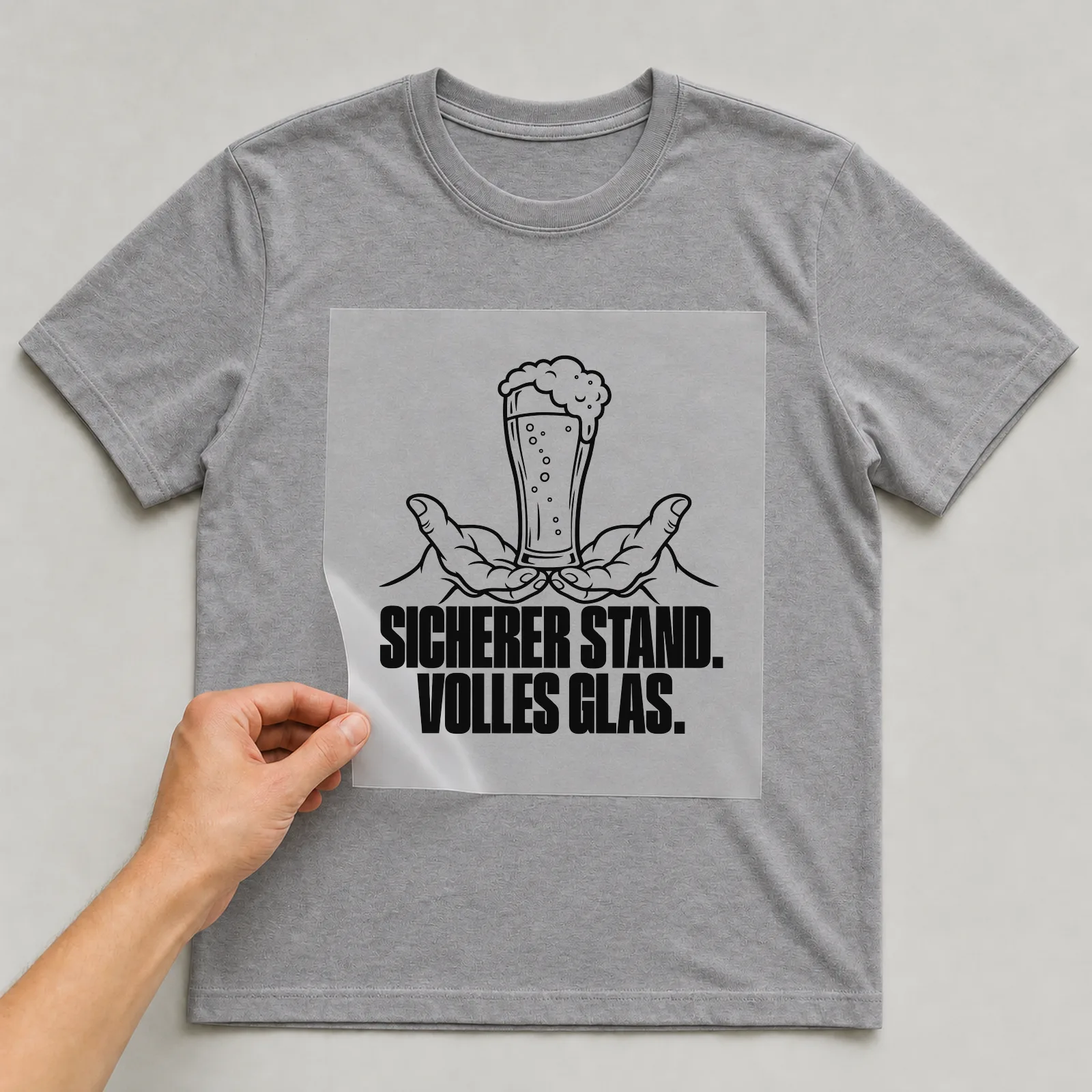 Bügelbild „Sicherer stand. Volles Glas.“ auf grauem T-Shirt, Folie wird aufgelegt