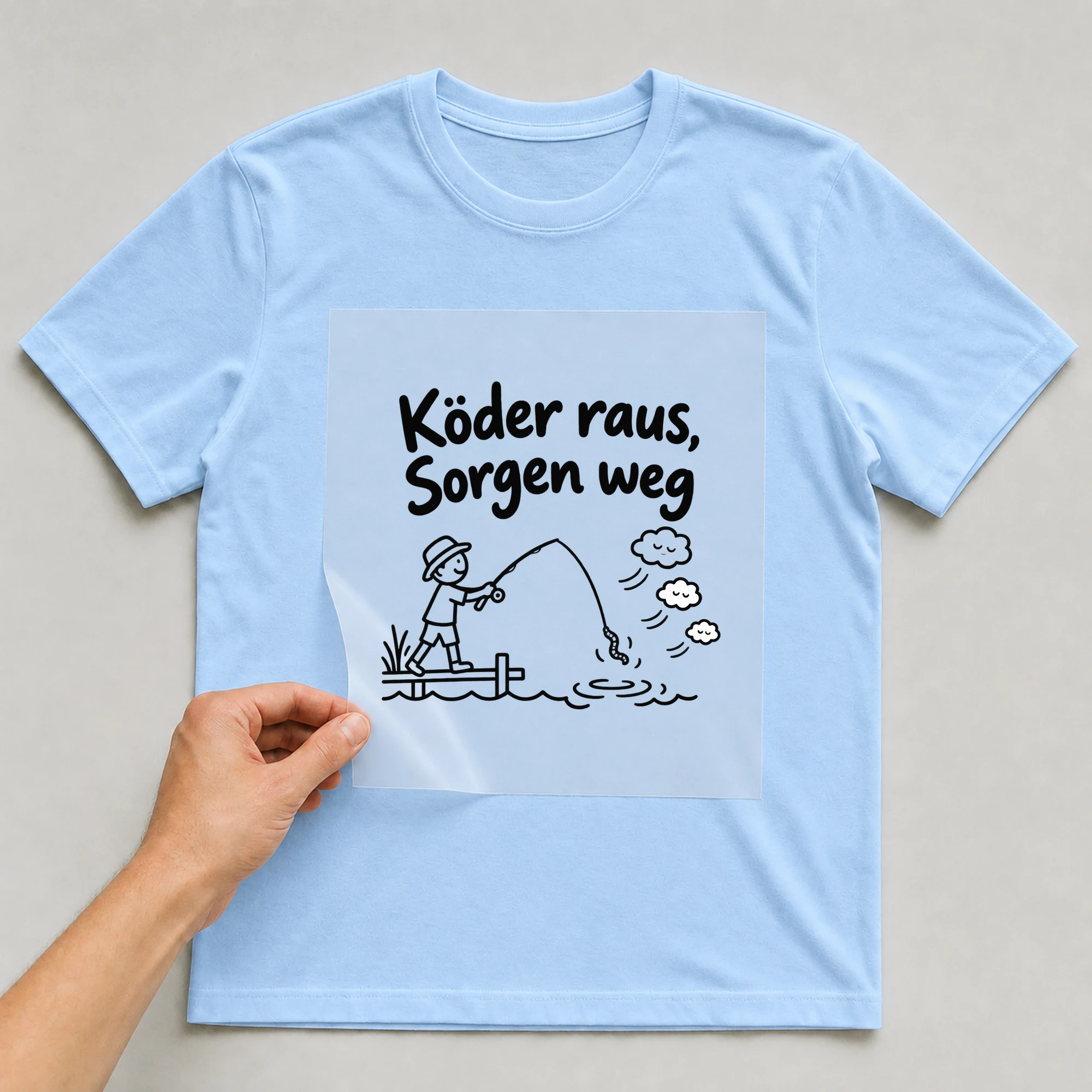 Bügelbild „Köder raus, Sorgen weg“ auf babyblauem T-Shirt, Folie wird aufgelegt