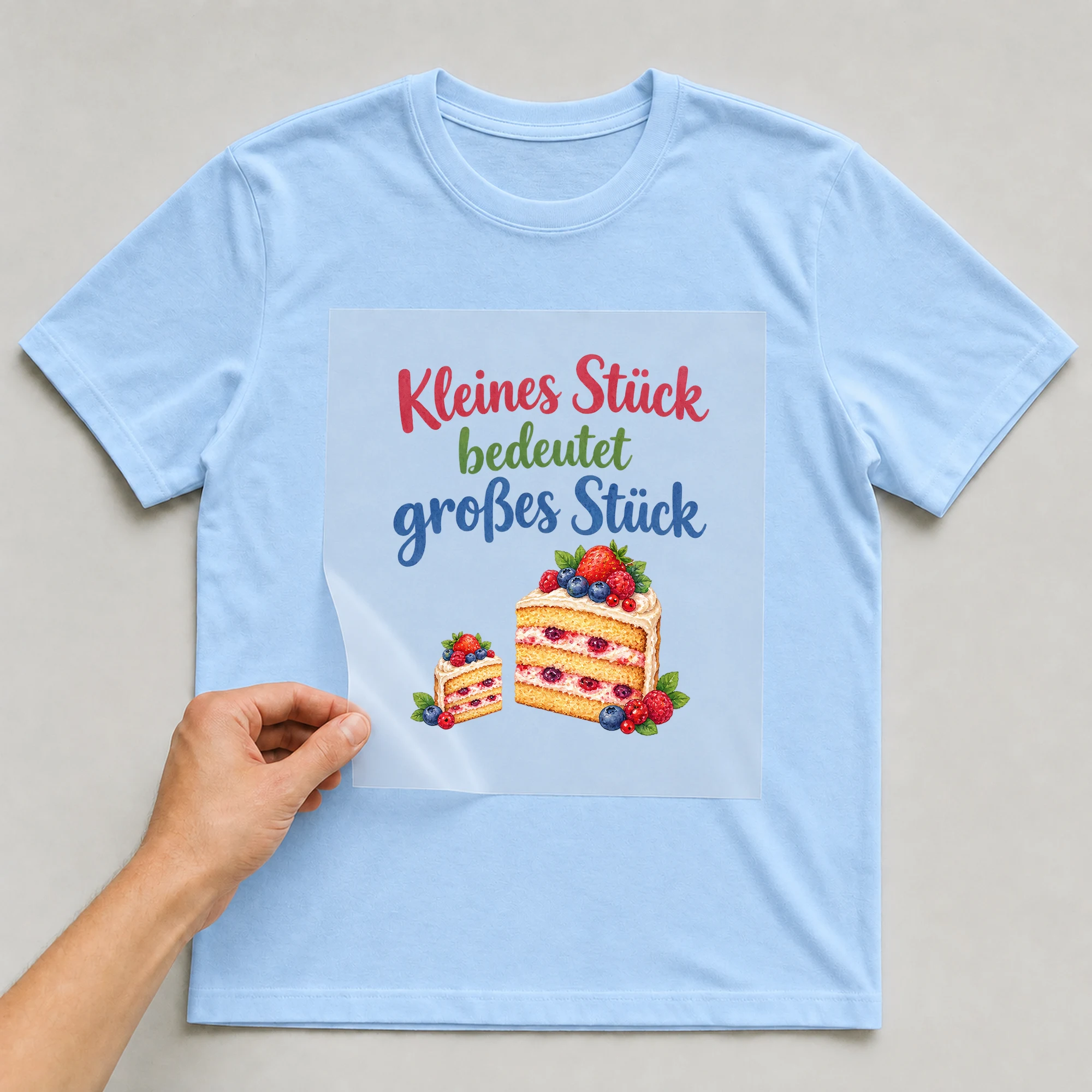 Bügelbild „Kleines Stück bedeutet großes Stück“ auf babyblauem T-Shirt, Folie wird aufgelegt