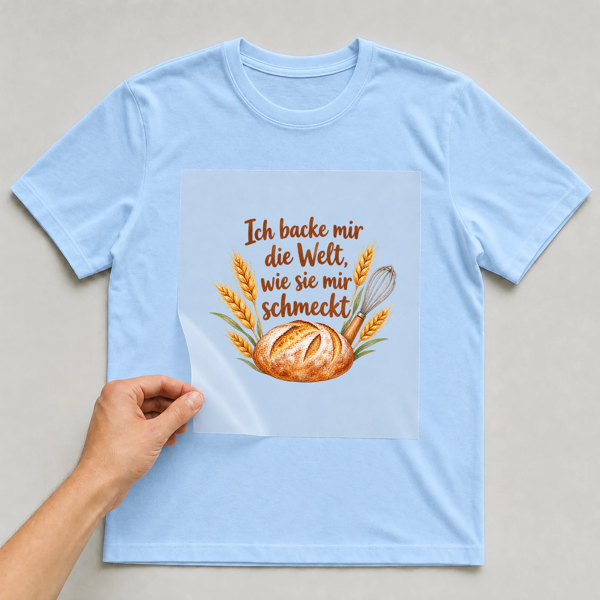 Bügelbild „Ich backe mir die Welt, wie sie mir schmeckt“ auf babyblauem T-Shirt, Folie wird aufgelegt