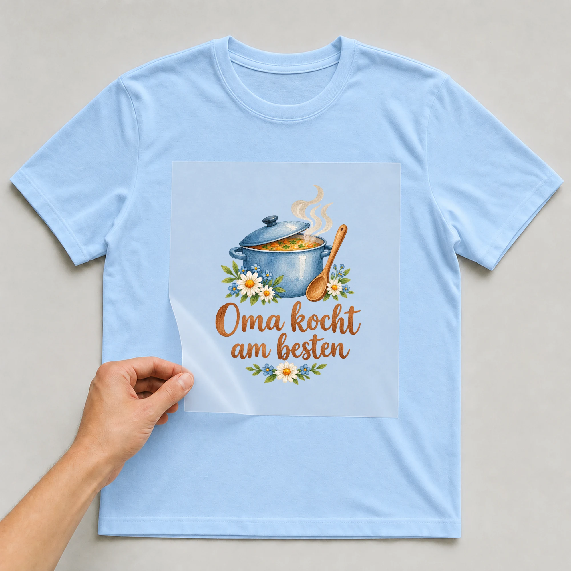 Bügelbild „Oma kocht am besten“ auf babyblauem T-Shirt, Folie wird aufgelegt