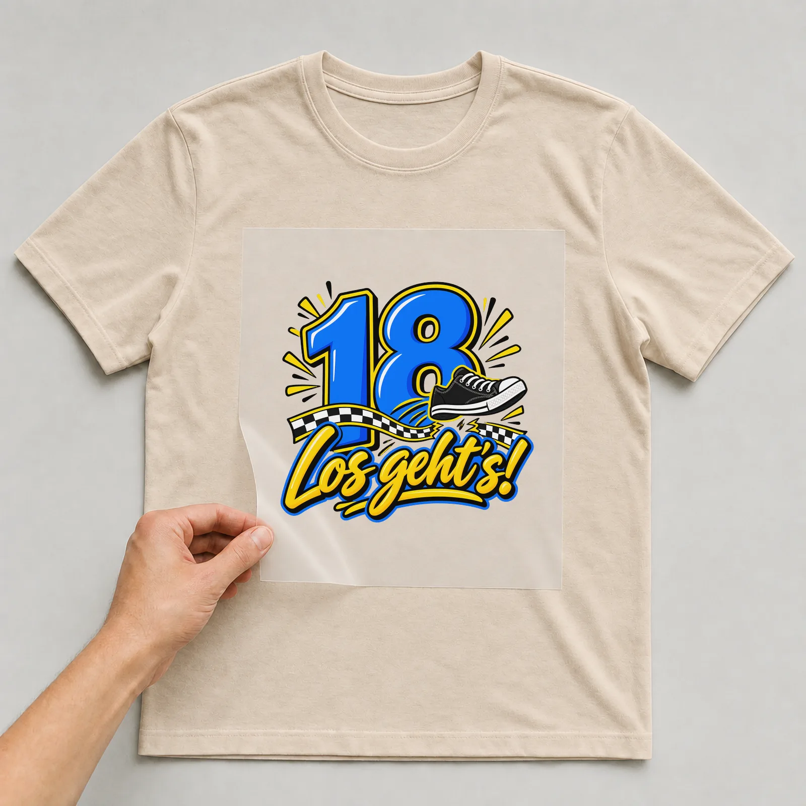 Bügelbild „18 · Los geht’s!“ auf beigem T-Shirt, Folie wird aufgelegt