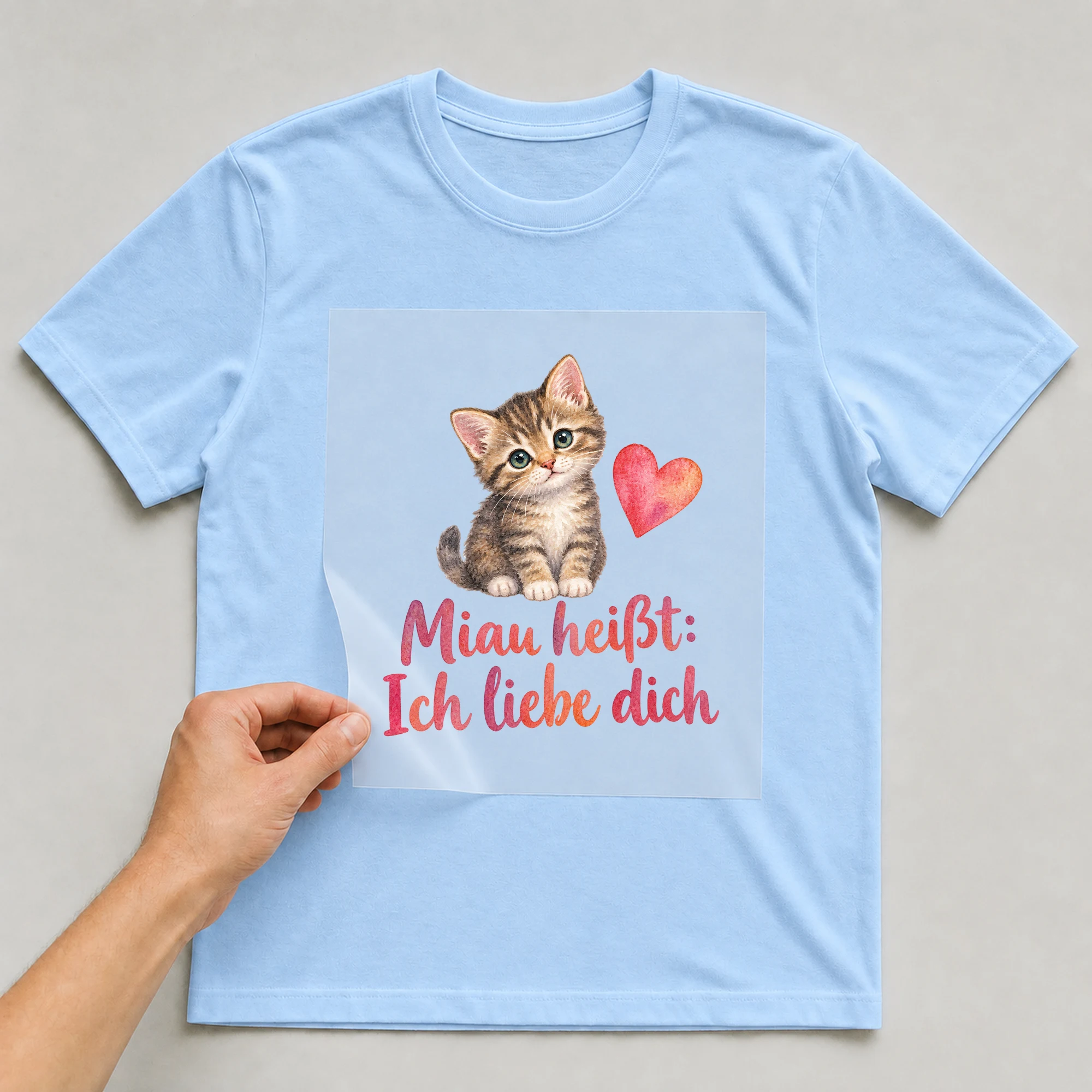 Bügelbild „Miau heißt: Ich liebe dich“ auf babyblauem T-Shirt, Folie wird aufgelegt