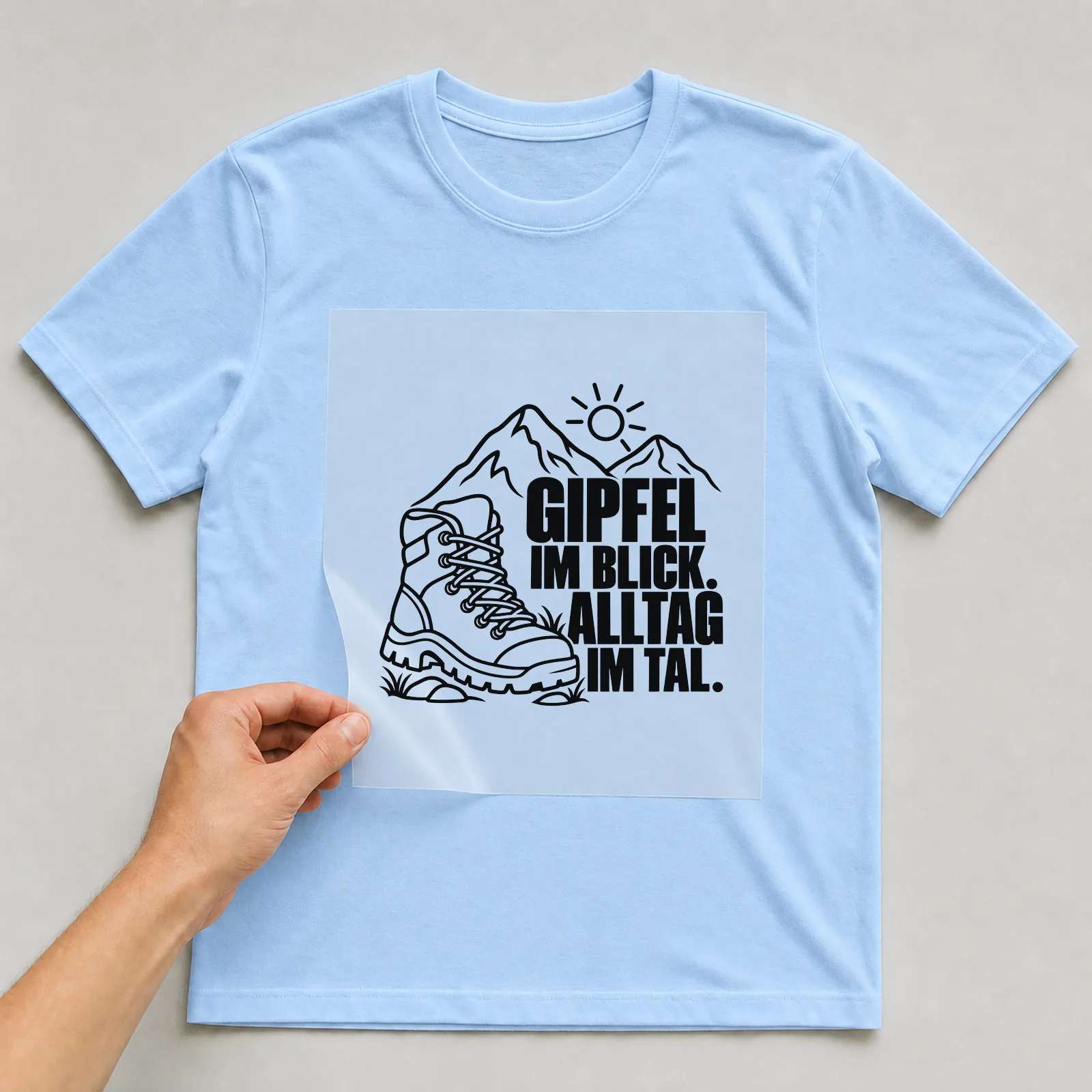 Bügelbild „Gipfel im Blick. Alltag im Tal.“ auf hellblauem T-Shirt, Folie wird aufgelegt