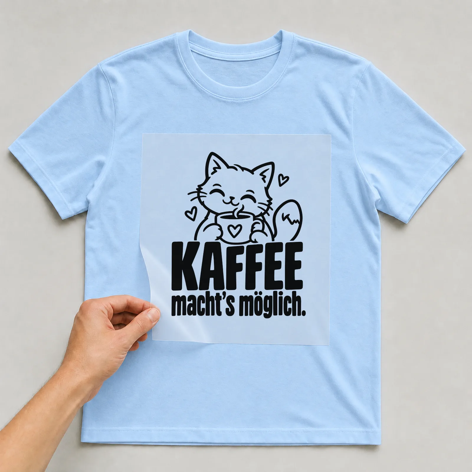 Bügelbild „Kaffee macht’s möglich“ auf hellblauem T-Shirt, Folie wird aufgelegt