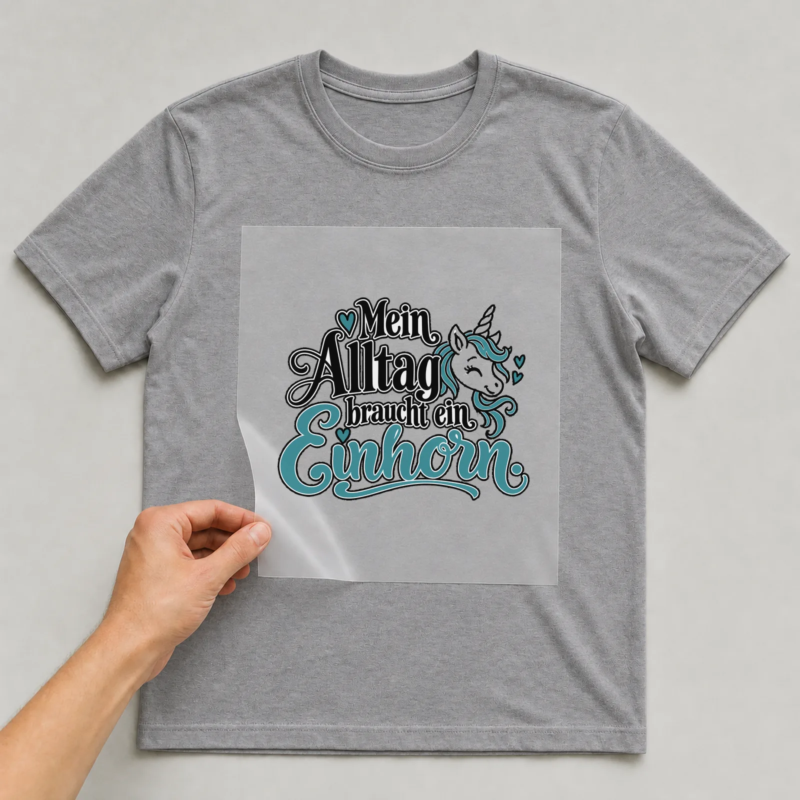 Bügelbild „Mein Alltag braucht ein Einhorn“ auf grauem T-Shirt, Folie wird aufgelegt