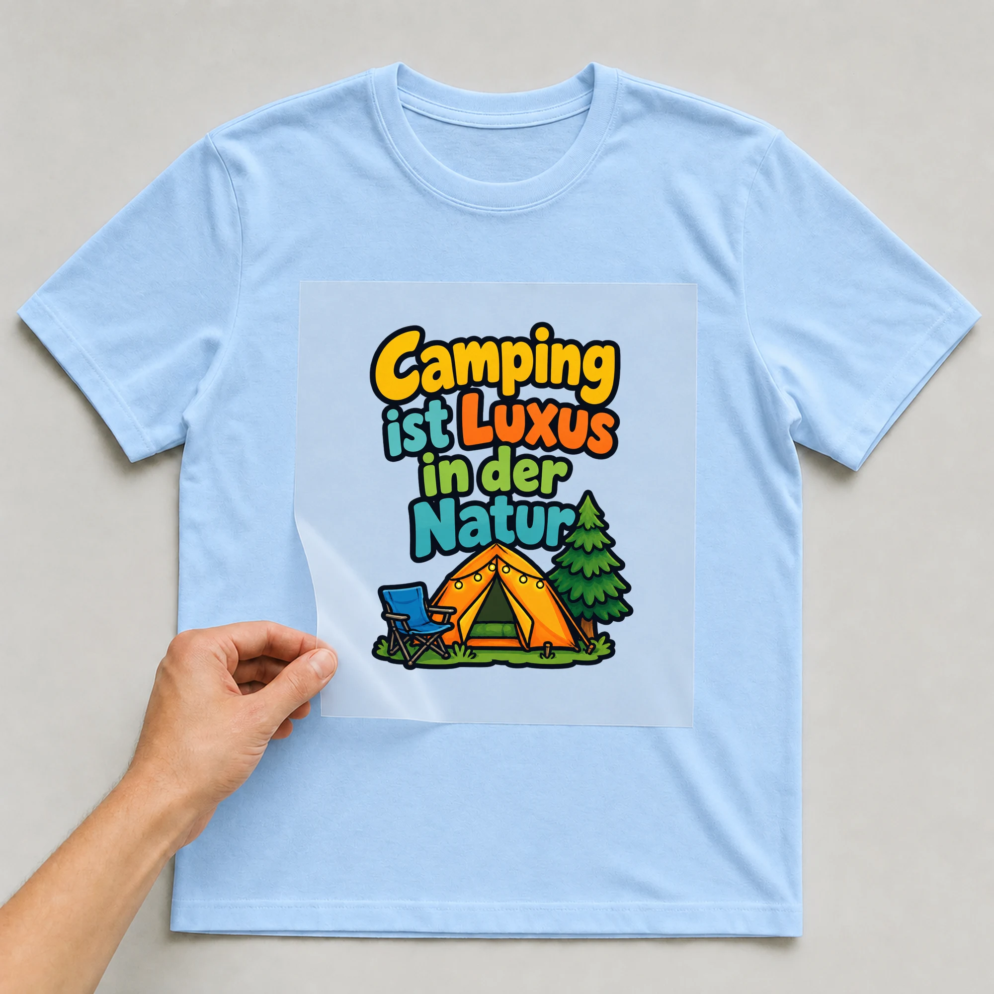 Bügelbild „Camping ist luxus in der natur“ auf babyblauem T-Shirt, Folie wird aufgelegt