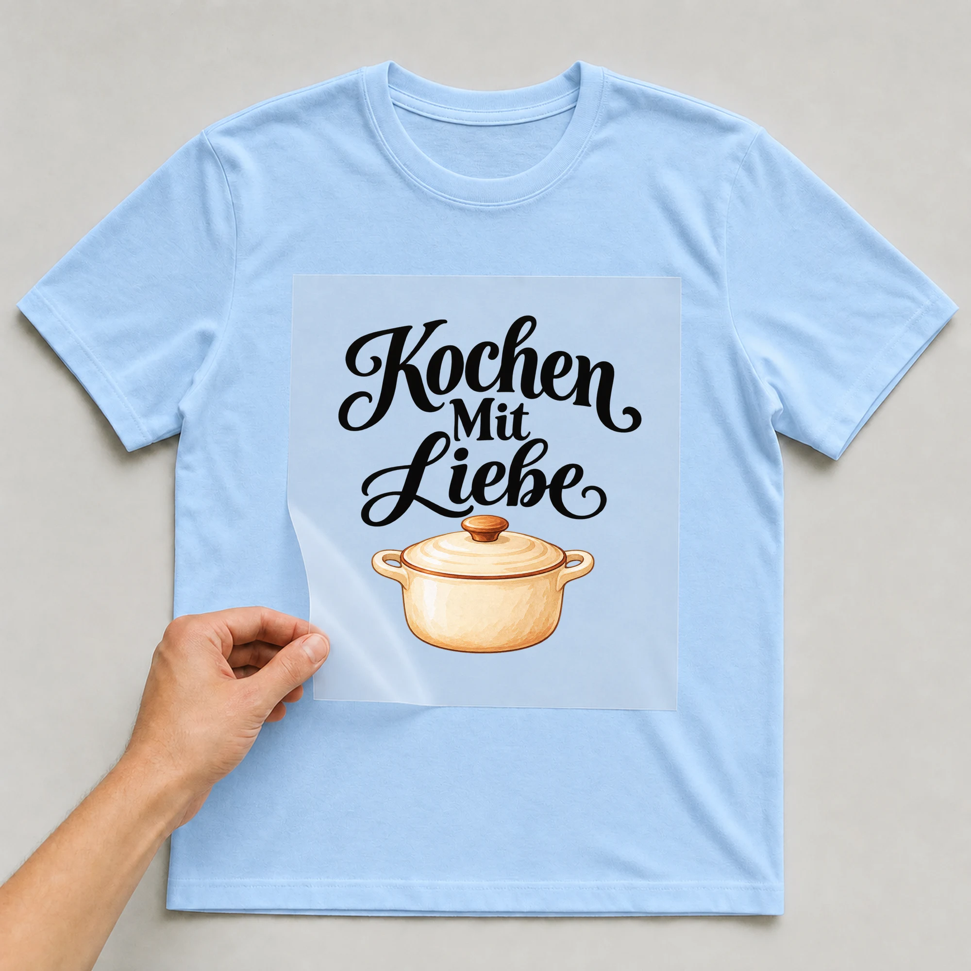 Bügelbild „Kochen Mit Liebe“ auf babyblauem T-Shirt, Folie wird aufgelegt