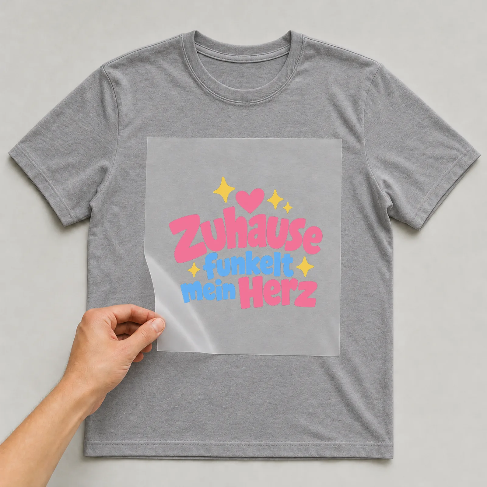 Bügelbild „Zuhause funkelt mein Herz“ auf grauem T-Shirt, Folie wird aufgelegt