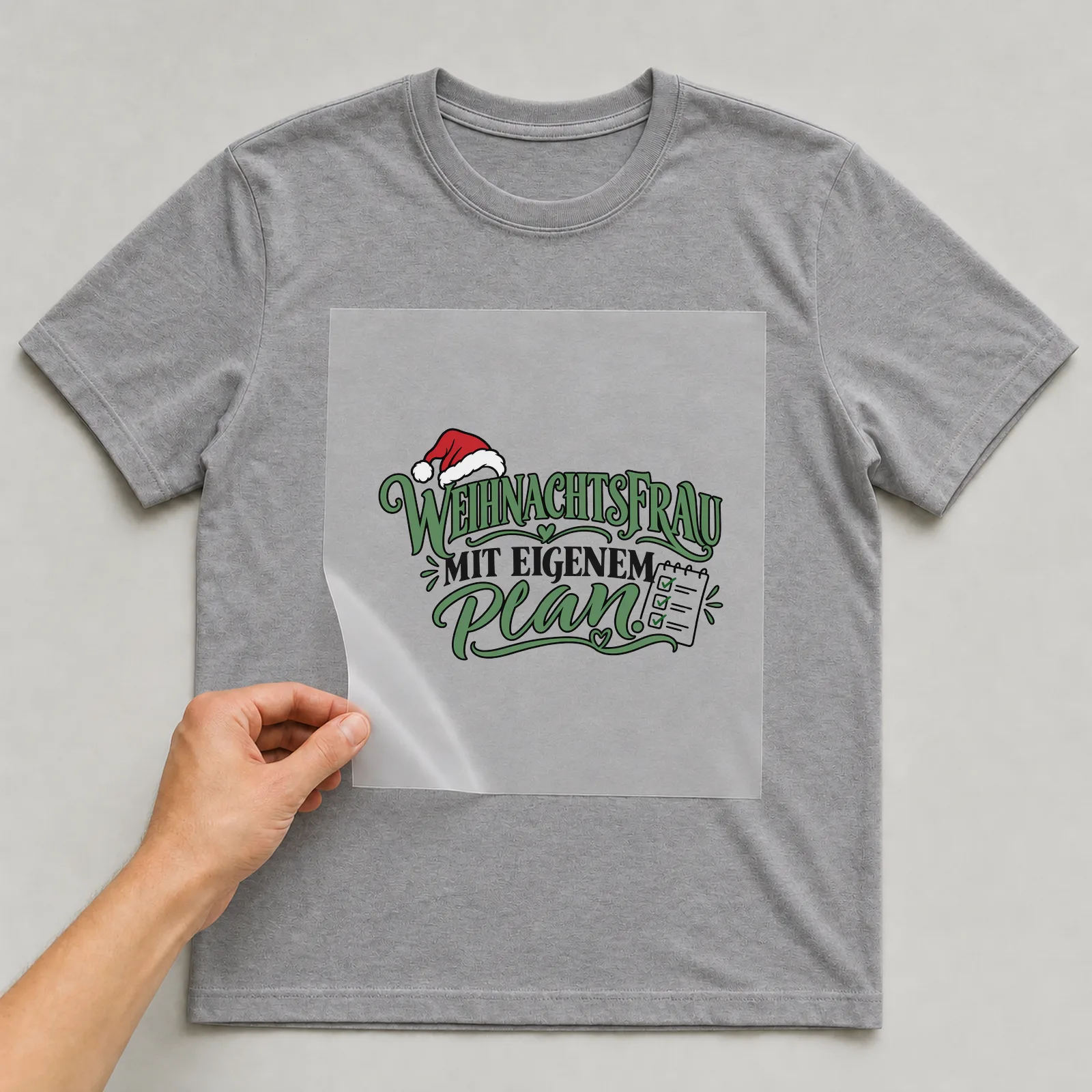 Bügelbild „Weihnachtsfrau mit eigenem Plan“ auf grauem T-Shirt, Folie wird aufgelegt