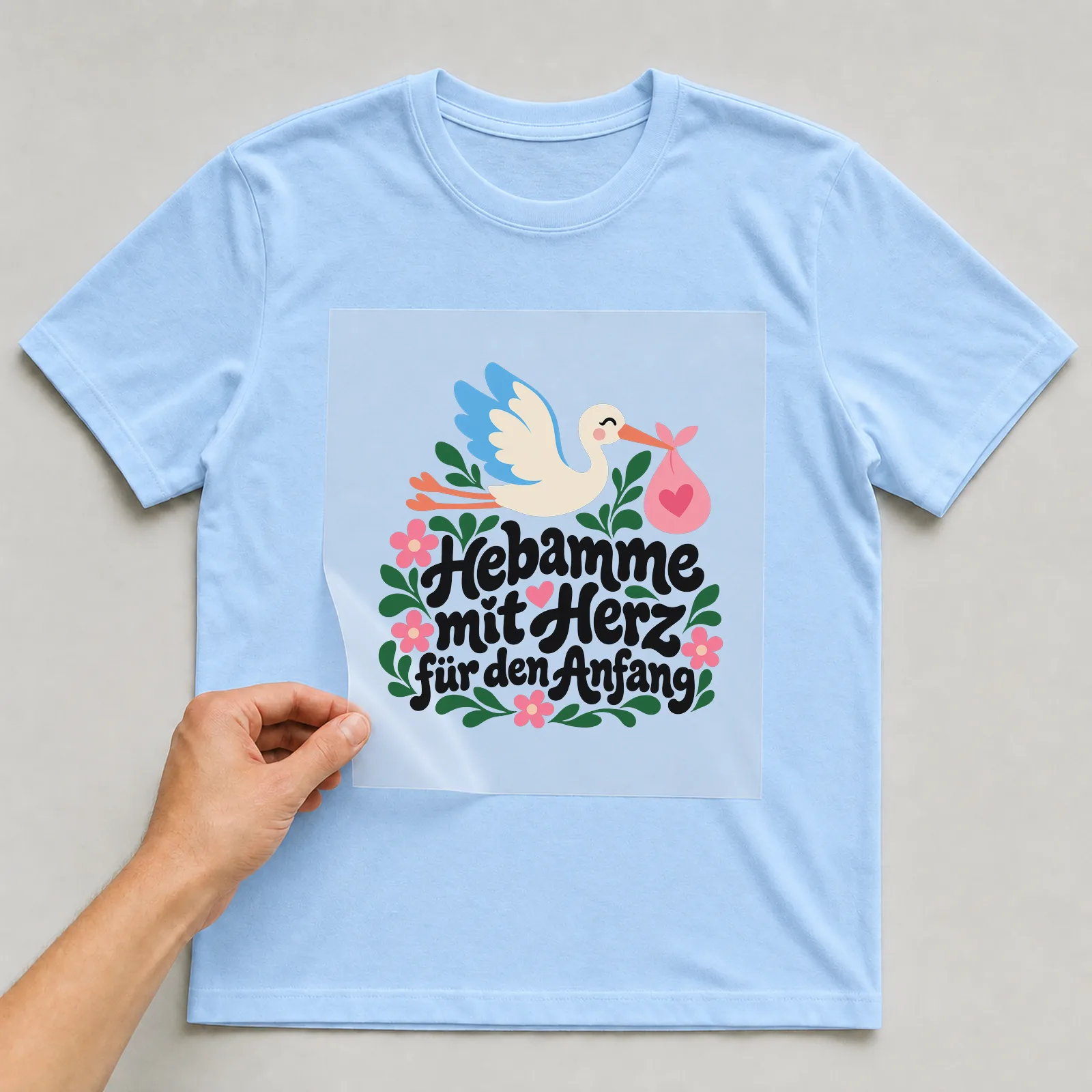 Bügelbild „Hebamme mit Herz für den Anfang“ auf hellblauem T-Shirt, Folie wird aufgelegt