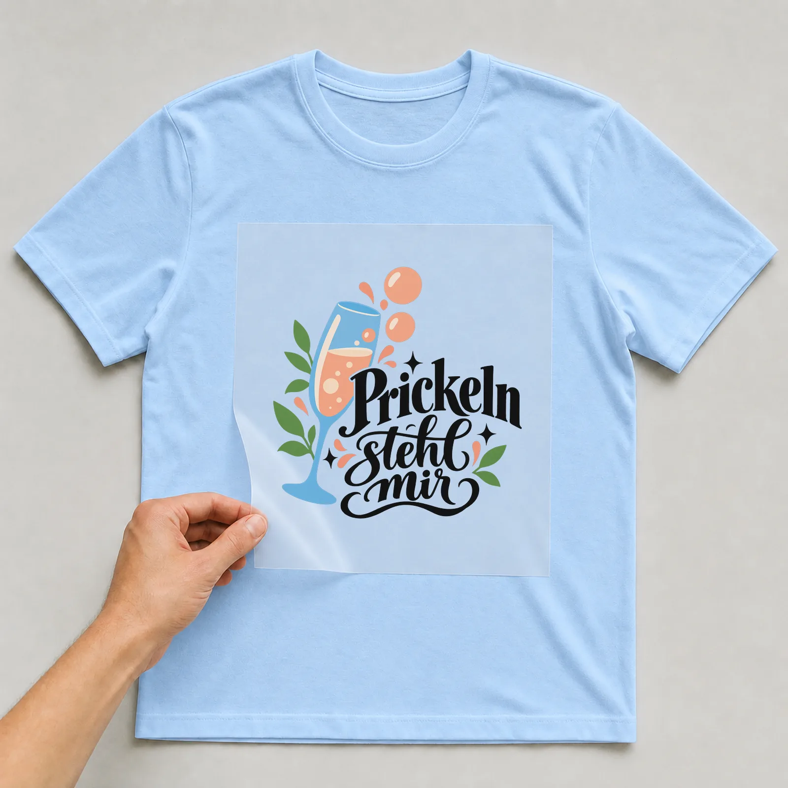 Bügelbild „Prickeln steht mir“ auf hellblauem T-Shirt, Folie wird aufgelegt
