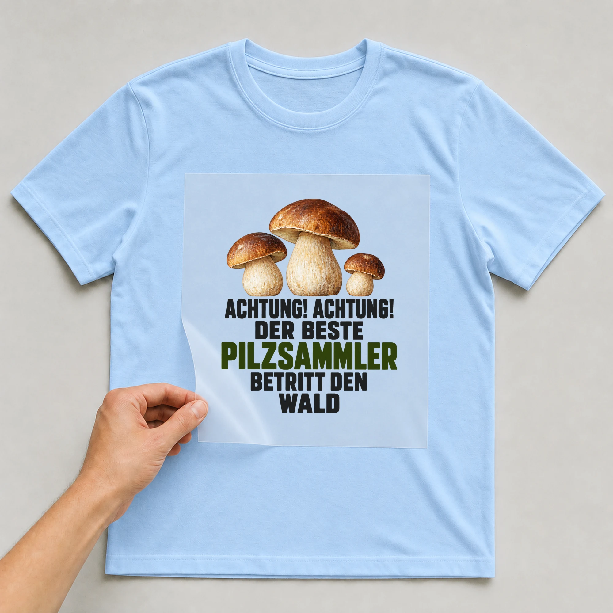 Bügelbild „Achtung! Achtung! Der beste Pilzsammler betritt den Wald“ auf babyblauem T-Shirt, Folie wird aufgelegt