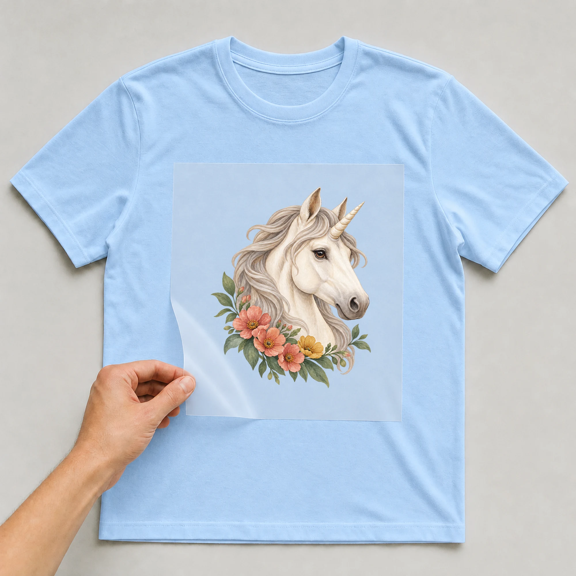 Bügelbild „Blumen Einhorn“ auf babyblauem T-Shirt, Folie wird aufgelegt