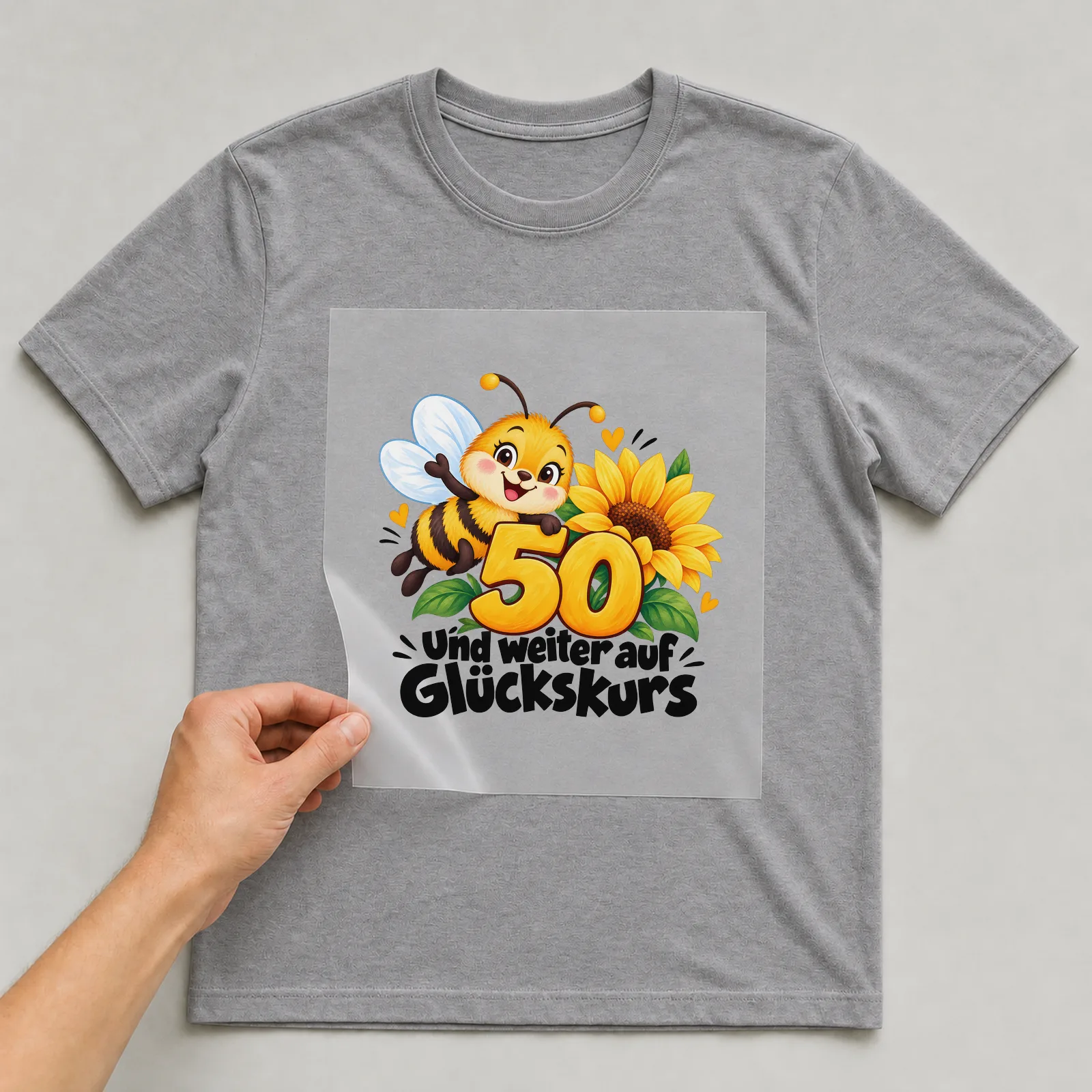 Bügelbild „50 und weiter auf Glückskurs“ auf grauem T-Shirt, Folie wird aufgelegt