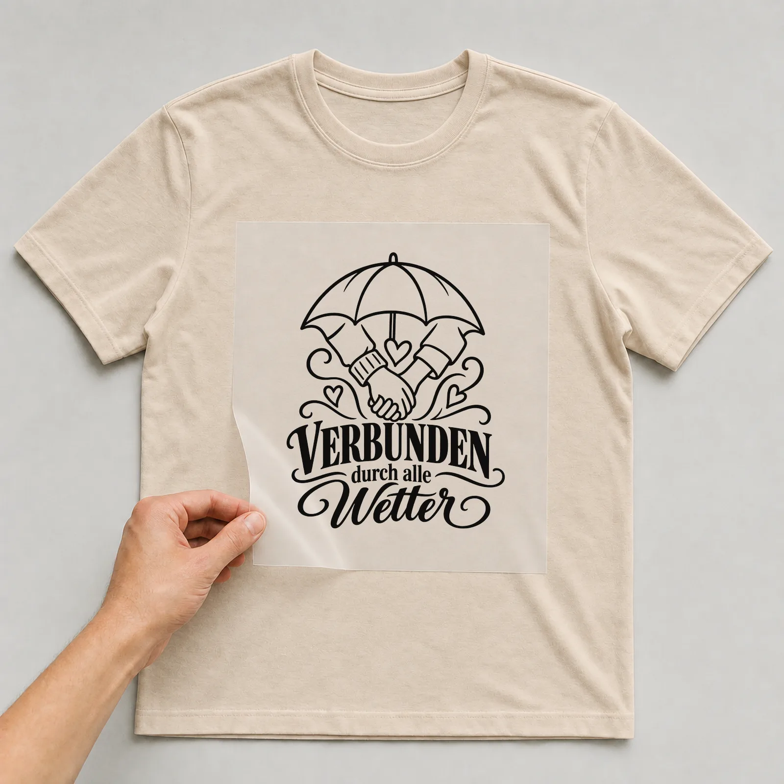 Bügelbild „VERBUNDEN durch alle Wetter“ auf beigem T-Shirt, Folie wird aufgelegt