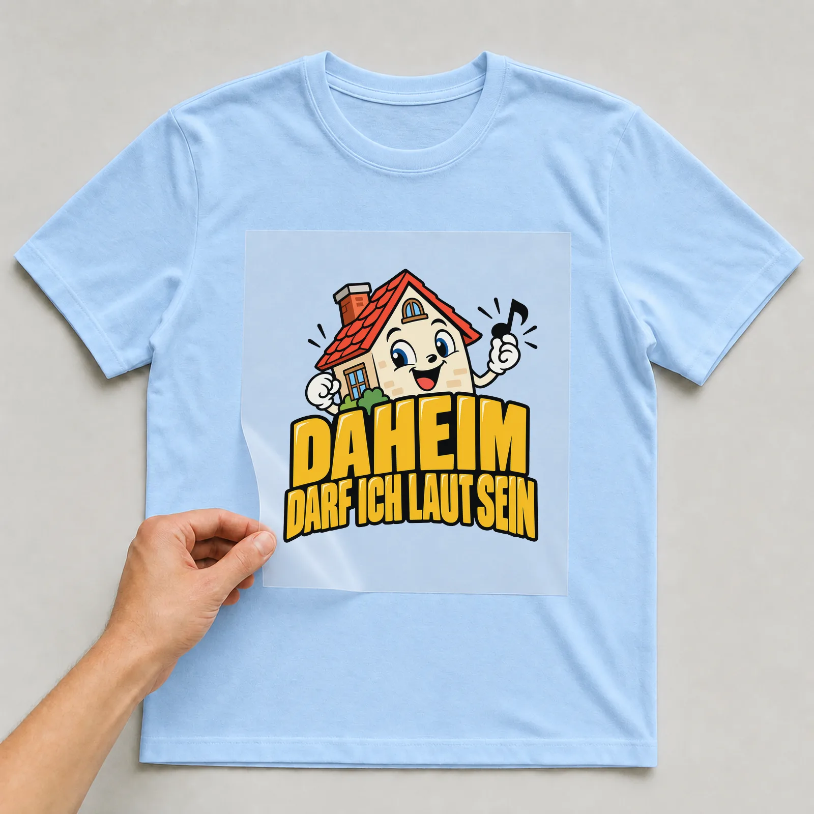 Bügelbild „Daheim darf ich laut sein“ auf hellblauem T-Shirt, Folie wird aufgelegt