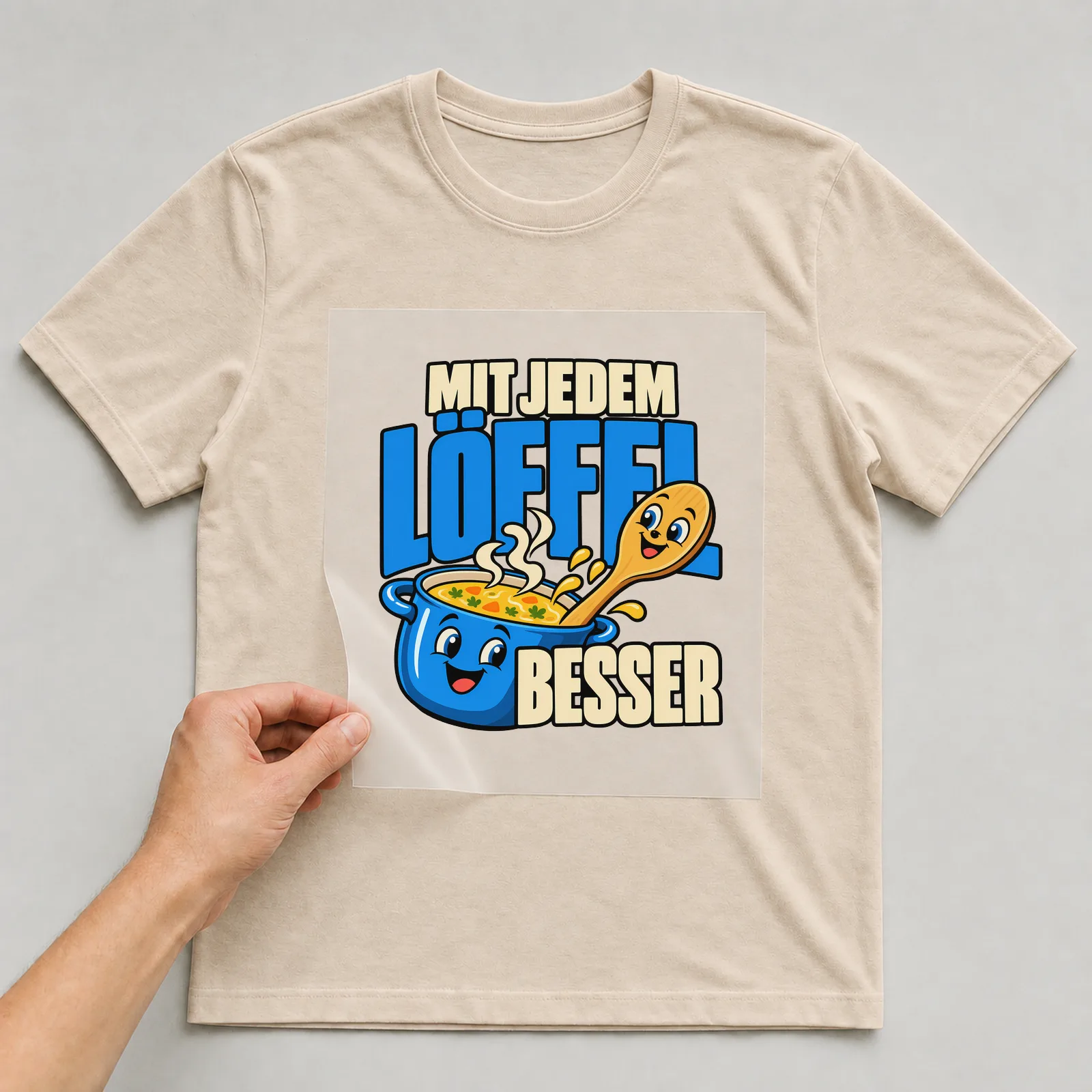 Bügelbild „Mit jedem Löffel besser“ auf beigem T-Shirt, Folie wird aufgelegt
