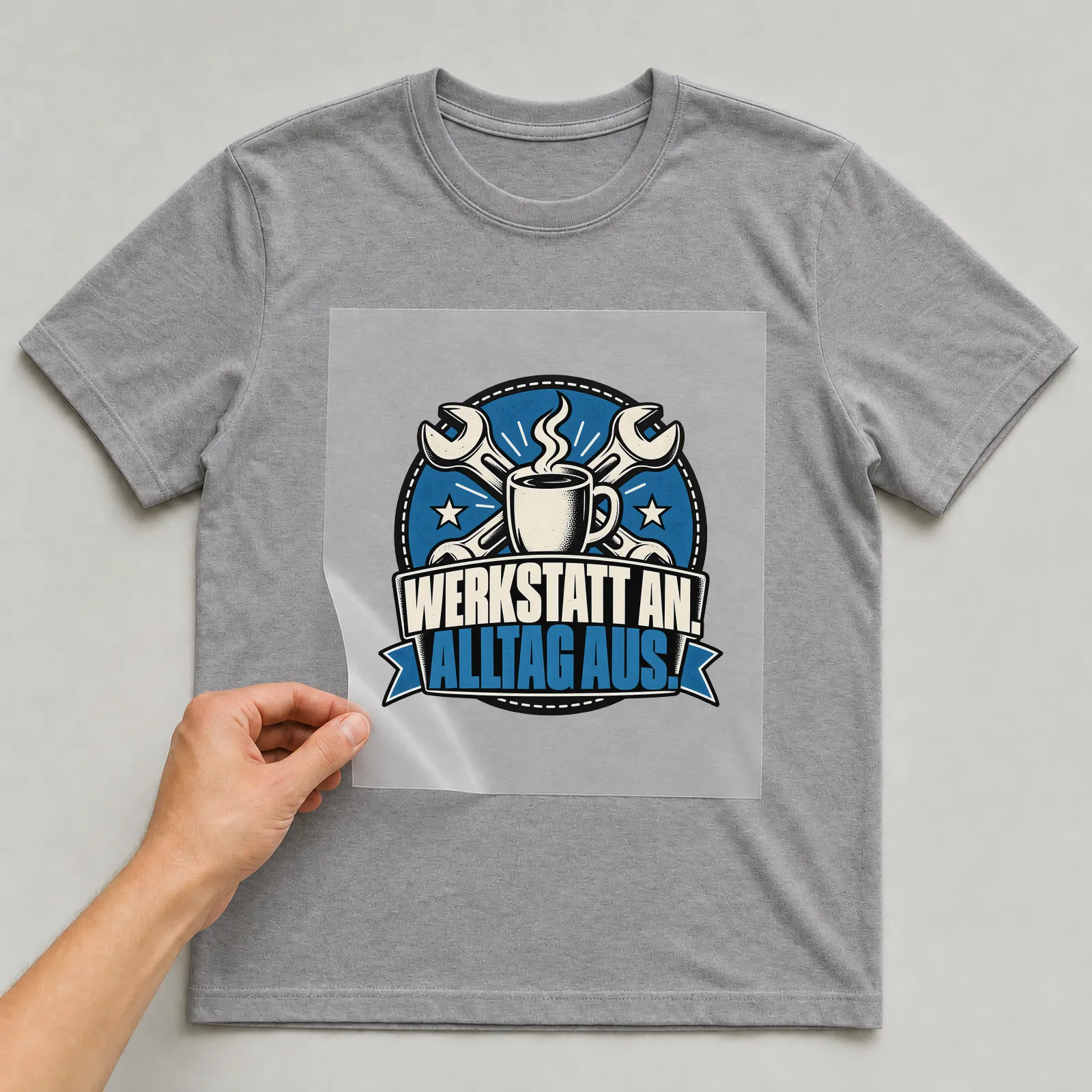 Bügelbild „Werkstatt an. Alltag aus.“ auf grauem T-Shirt, Folie wird aufgelegt