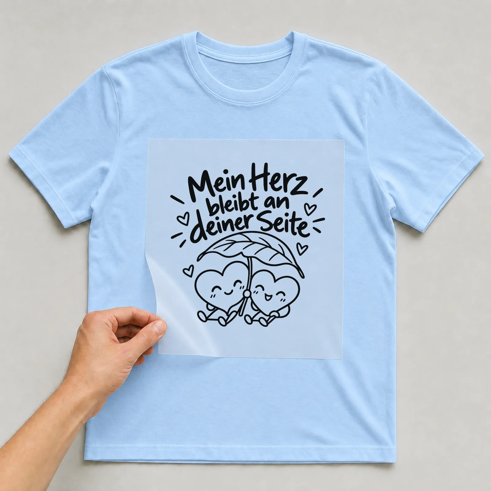 Bügelbild „Mein Herz bleibt an deiner Seite“ auf hellblauem T-Shirt, Folie wird aufgelegt