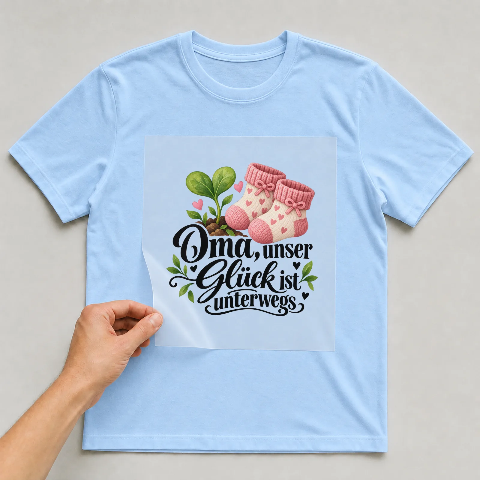 Bügelbild „Oma, unser Glück ist unterwegs“ auf hellblauem T-Shirt, Folie wird aufgelegt