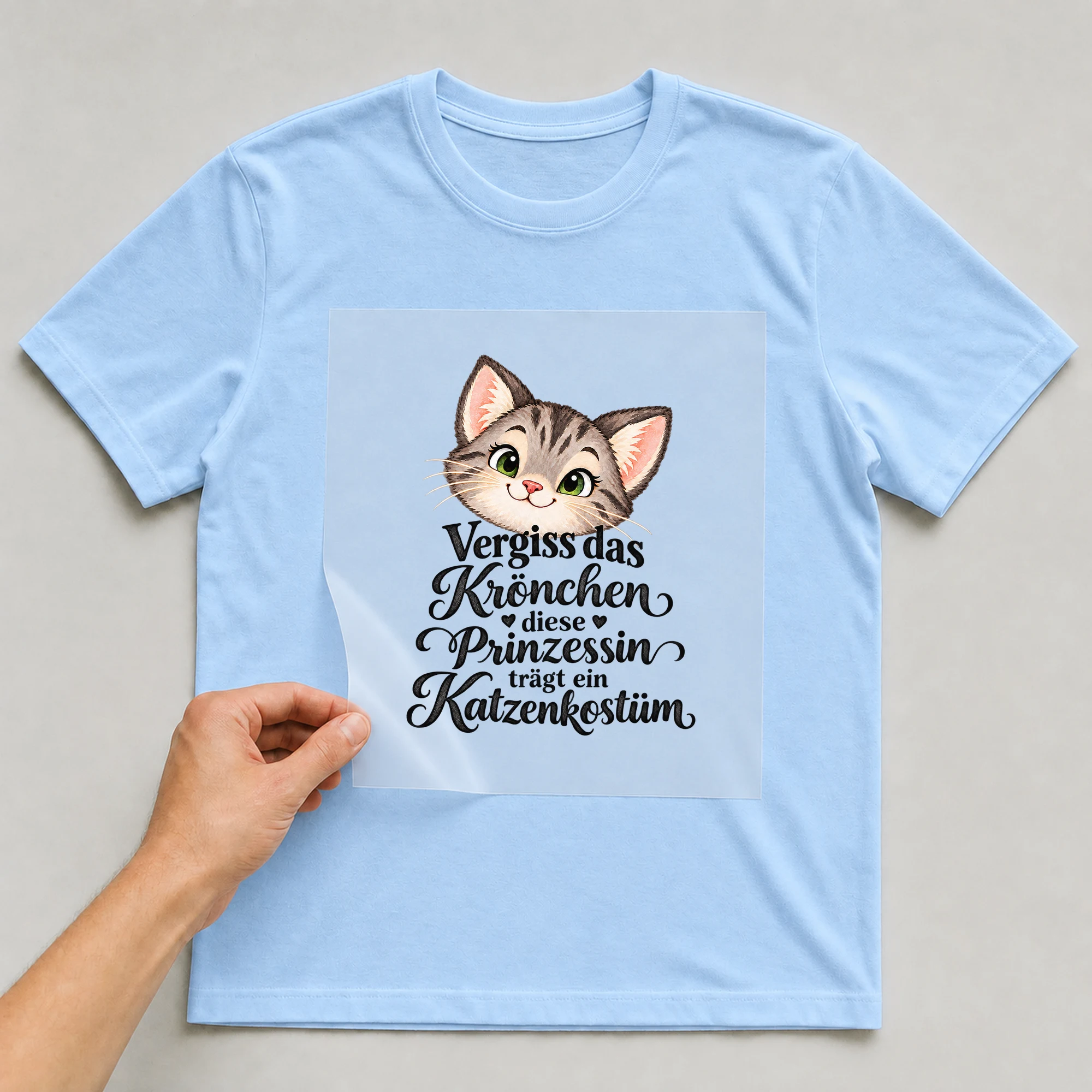 Bügelbild „Vergiss das Krönchen diese Prinzessin trägt ein Katzenkostüm“ auf babyblauem T-Shirt, Folie wird aufgelegt
