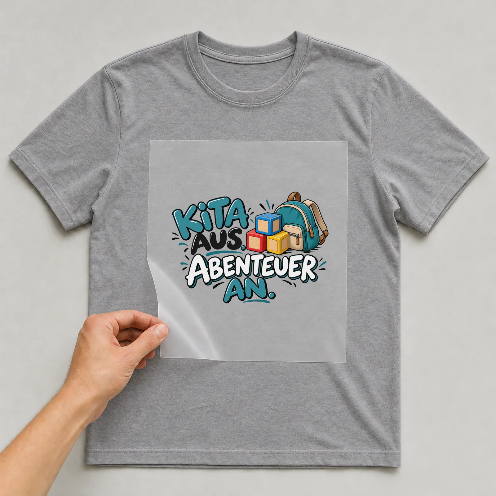 Bügelbild „Kita aus. Abenteuer an.“ auf grauem T-Shirt, Folie wird aufgelegt