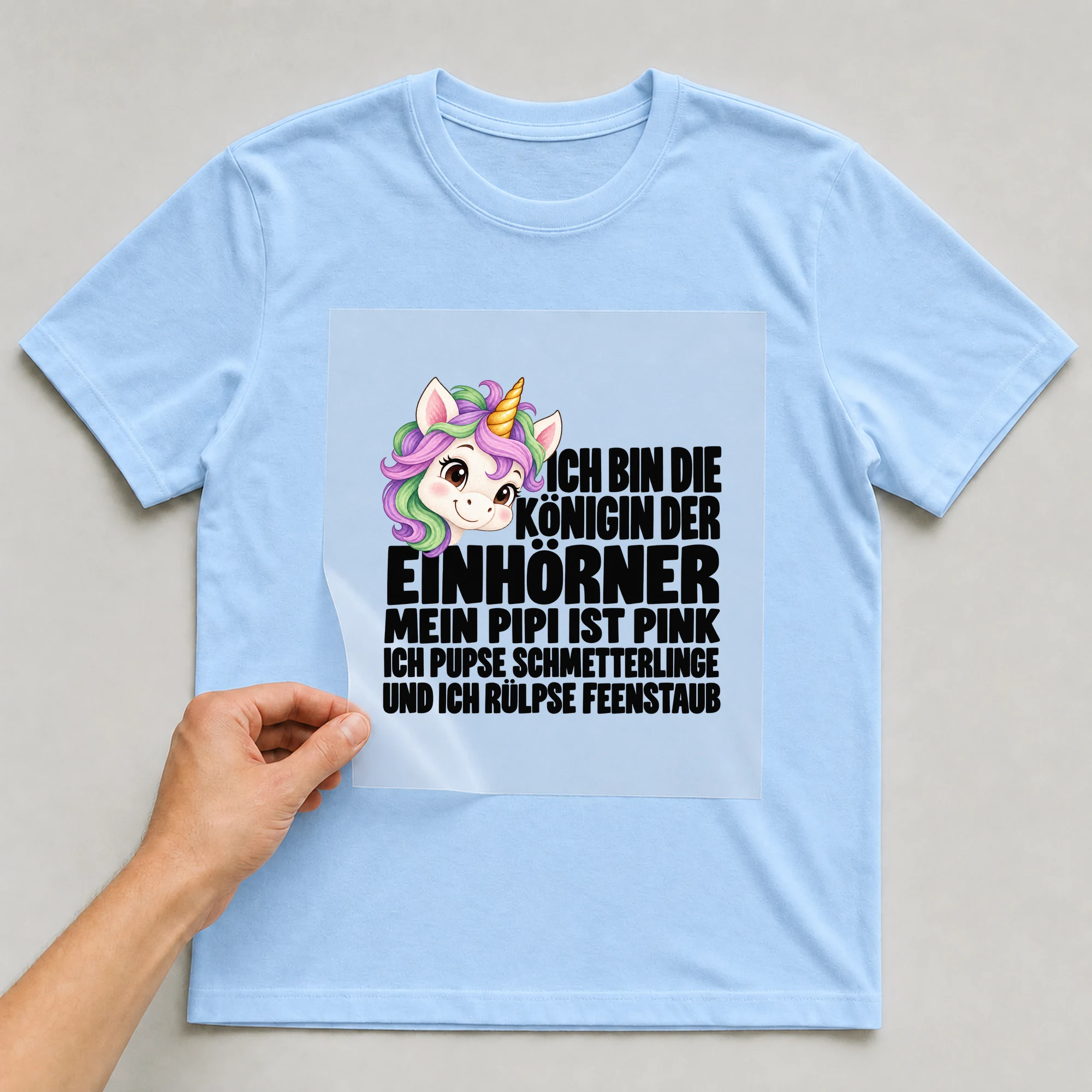 Bügelbild „Ich Bin Die Königin Der Einhörner Mein Pipi Ist Pink Ich Pupse Schmetterlinge“ auf babyblauem T-Shirt, Folie wird aufgelegt