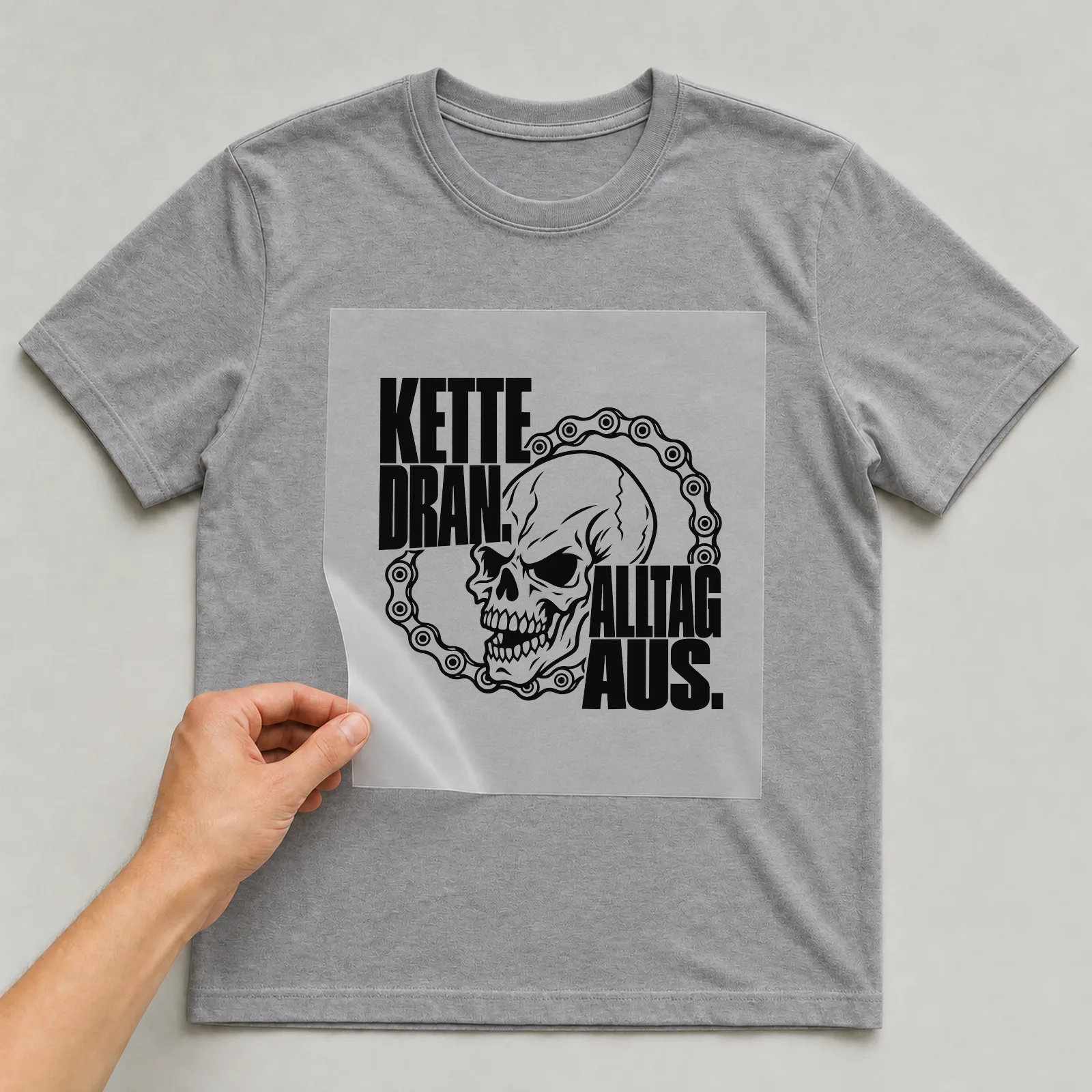 Bügelbild „Kette dran. Alltag aus.“ auf grauem T-Shirt, Folie wird aufgelegt