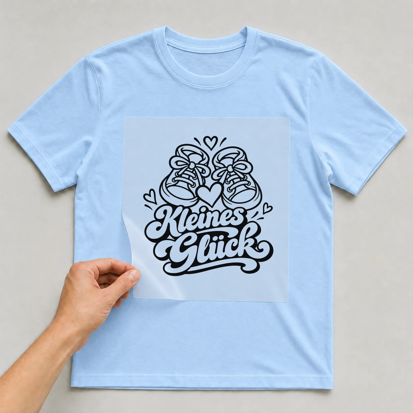 Bügelbild „Kleines Glück“ auf hellblauem T-Shirt, Folie wird aufgelegt