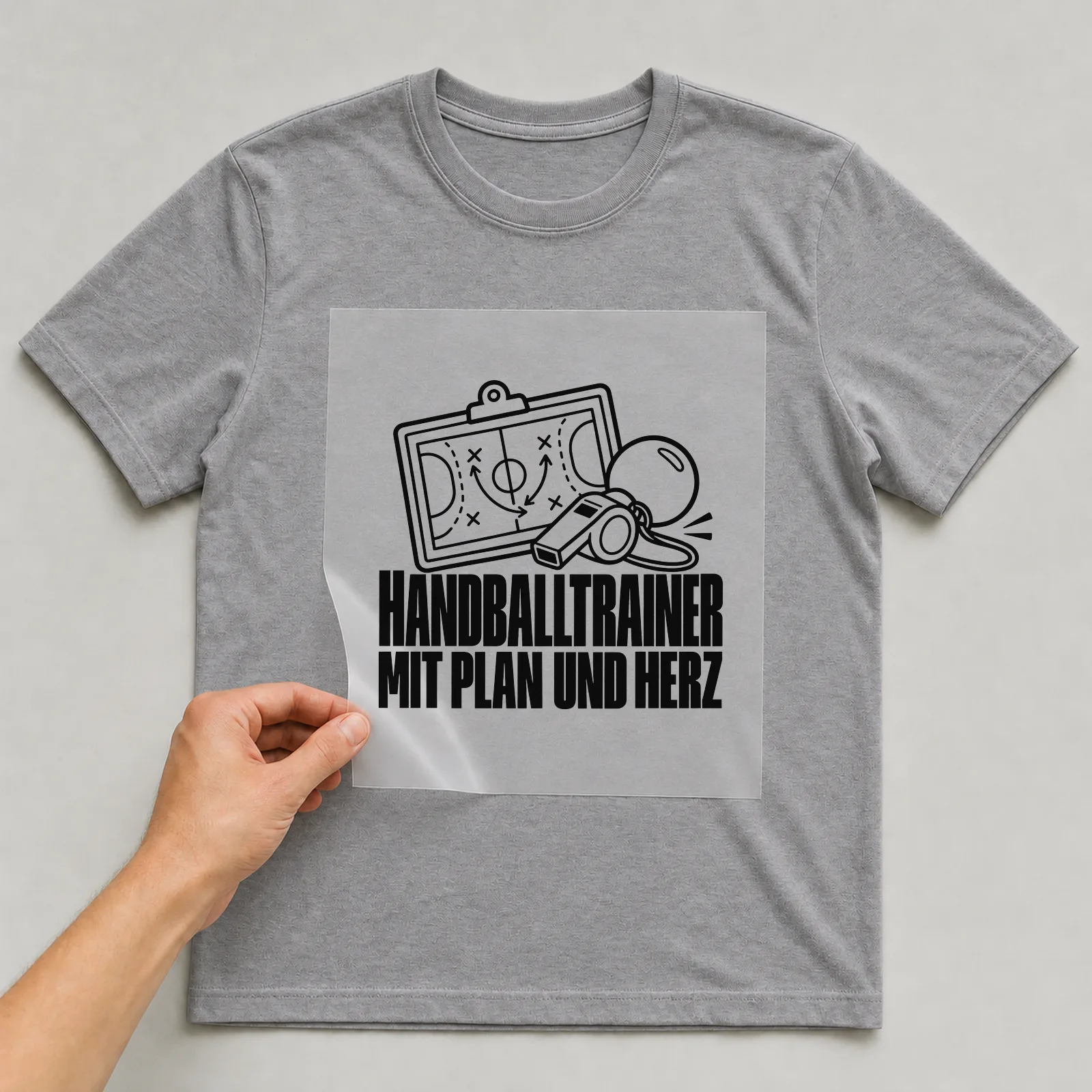 Bügelbild „Handballtrainer mit Plan und Herz“ auf grauem T-Shirt, Folie wird aufgelegt